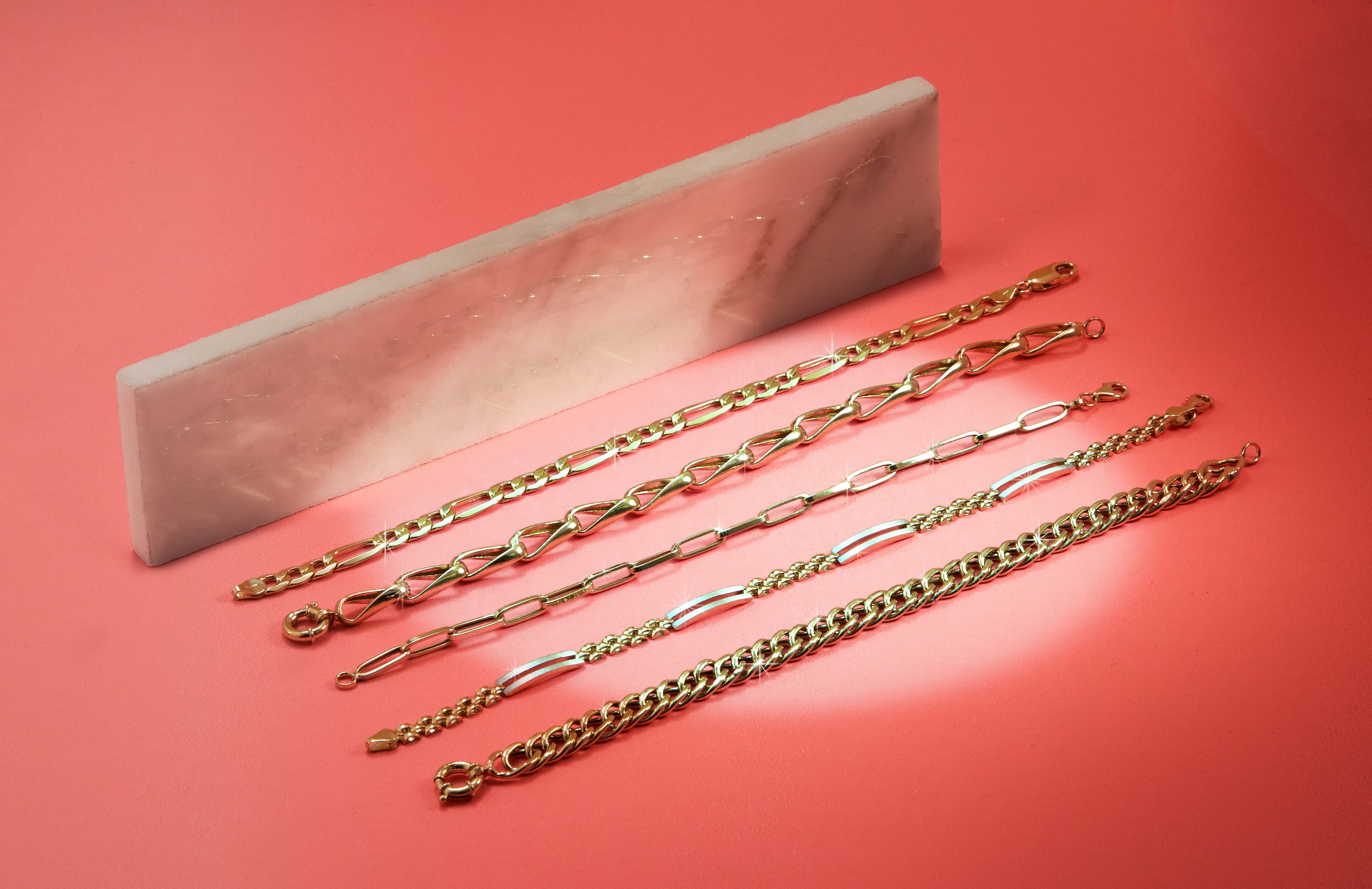 Several gold chains and bracelets laid out on a pink surface with a marble-like storage block in the background.