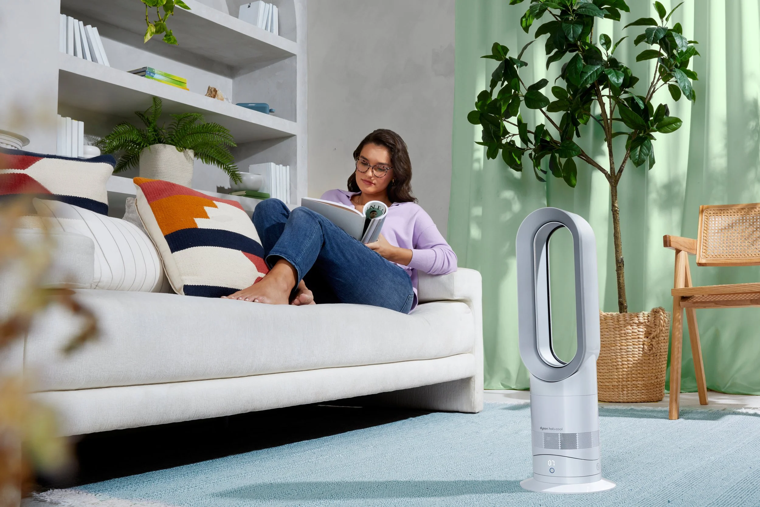 A woman sitting on a white couch reading a magazine in a living room with green curtains, plants, a wicker basket, and a Dyson bladeless fan.