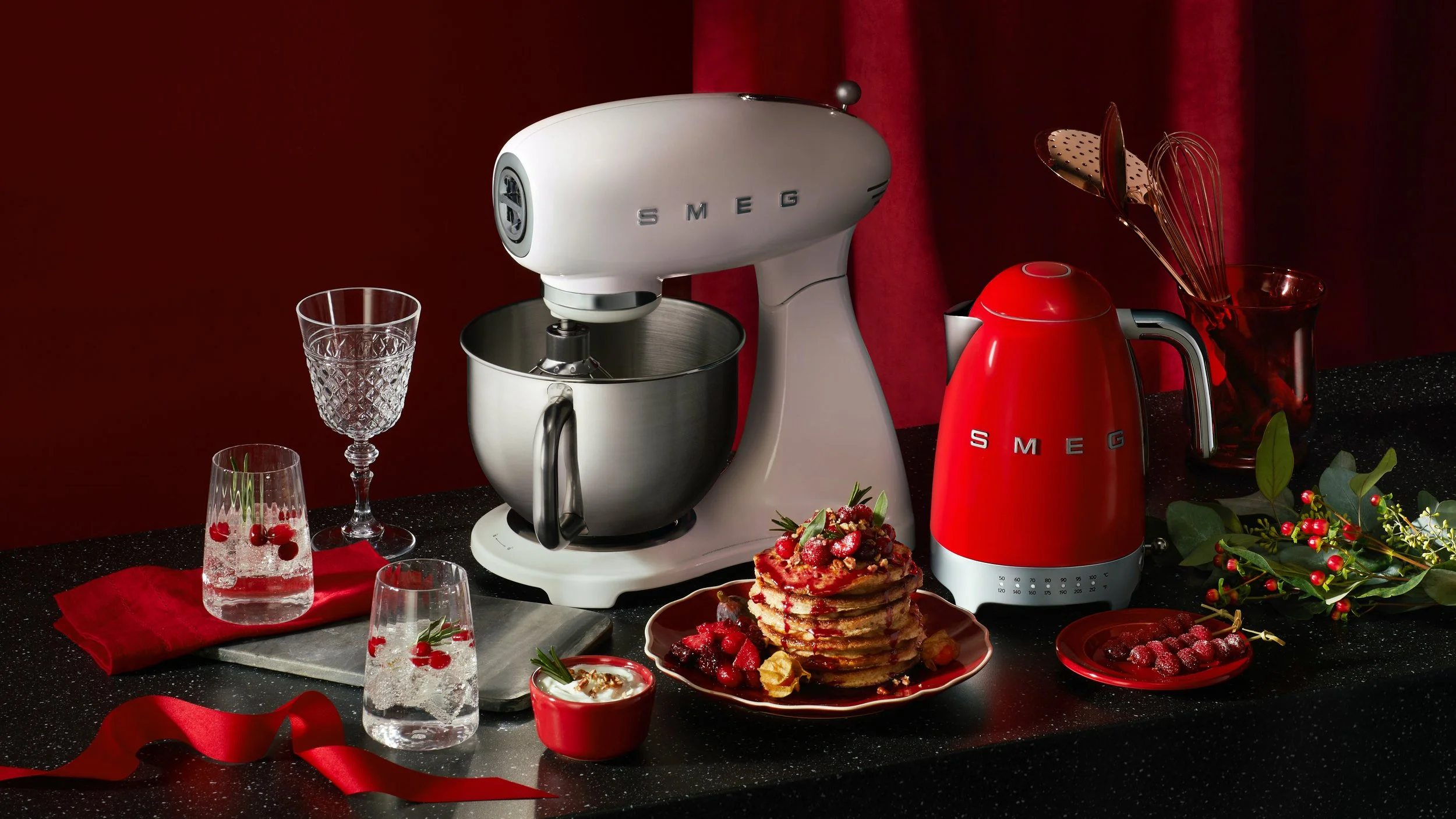 Holiday-themed kitchen countertop with red and white SMEG appliances, including a stand mixer and electric kettle, surrounded by glassware, pancakes topped with berries, and festive decorations.