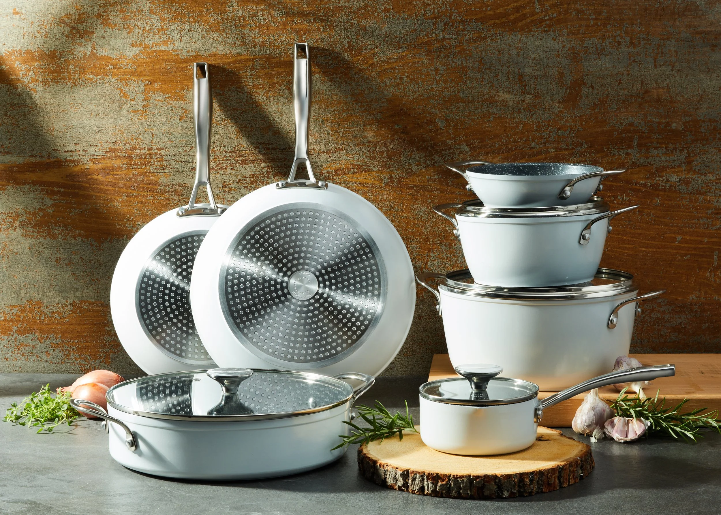 Set of white ceramic pots and pans with stainless steel handles, stacked on a wooden board and arranged on a kitchen countertop, with garlic, herbs, and onions around them.