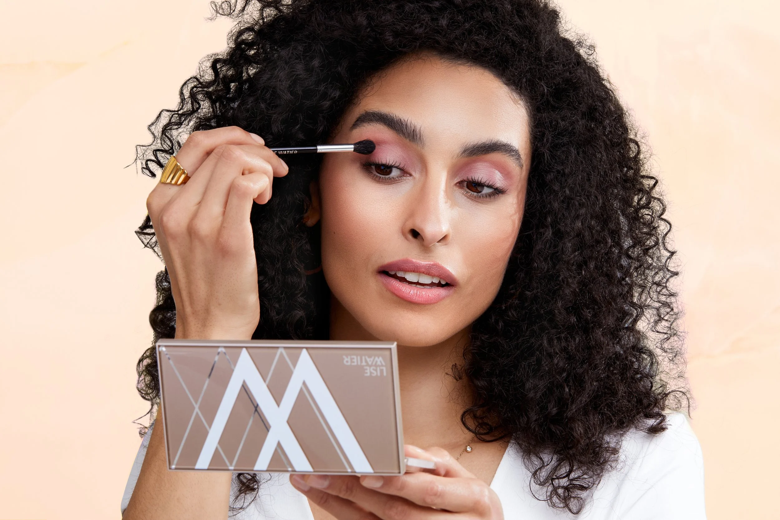 Woman with curly black hair applying eyeshadow while looking into a mirror.