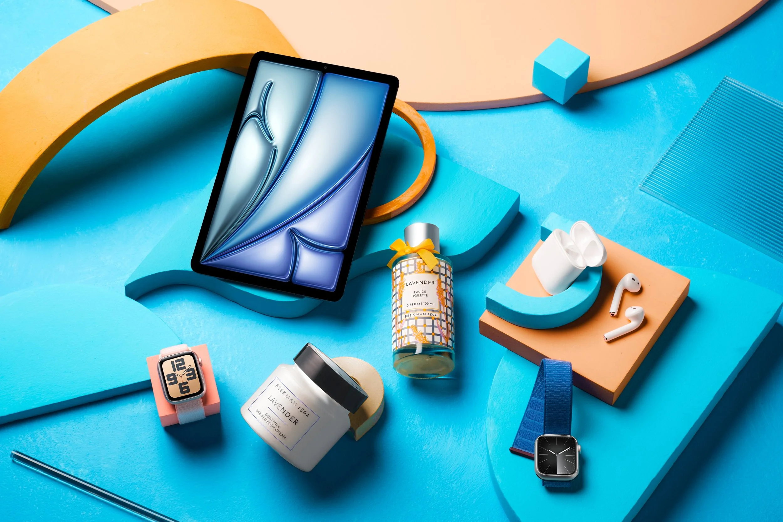 A tablet, smartwatch, fitness tracker, wireless earbuds, lavender perfume bottle, body cream, and decorative geometric shapes on a colorful abstract background.
