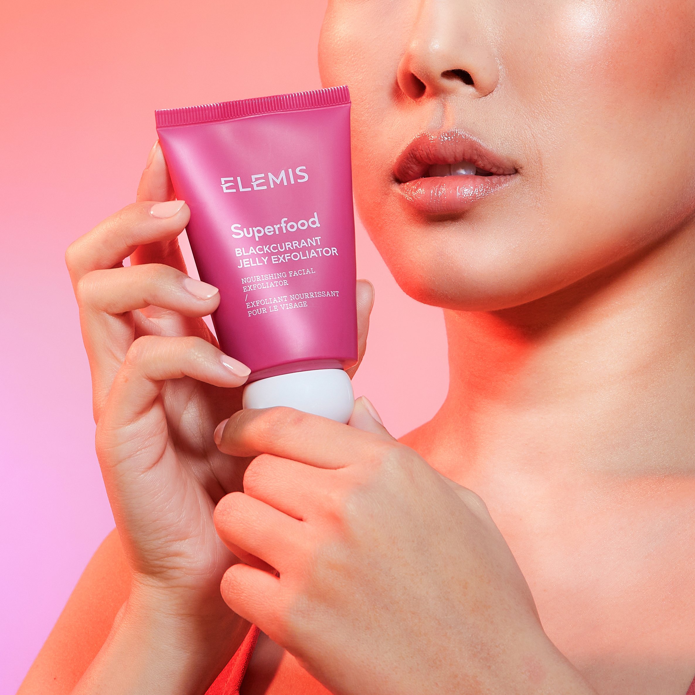 A woman holding a pink tube of Elemis Superfood Blackcurrant Jelly Exfoliator near her face, with a pink background.