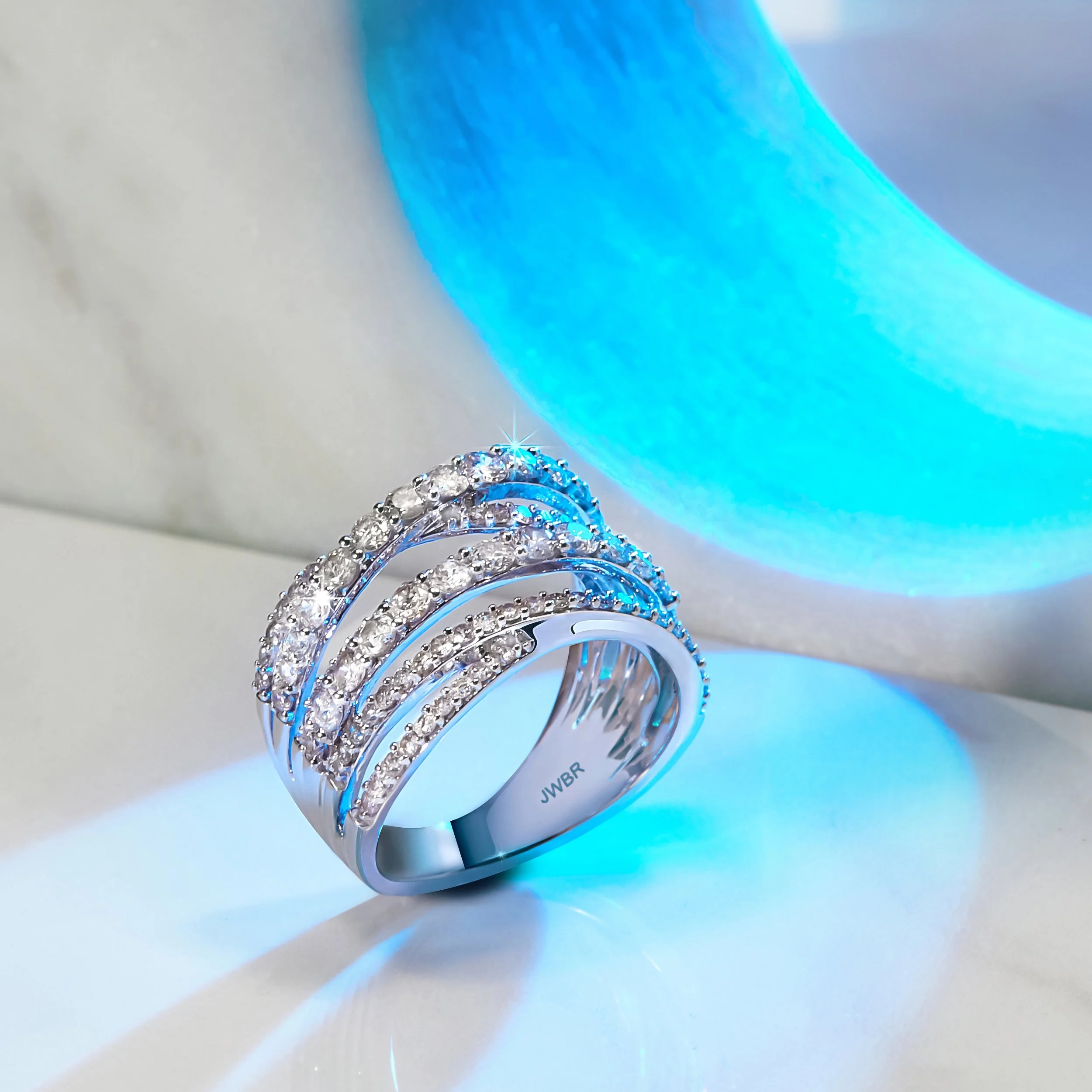 A silver ring with multiple curved rows of small diamonds, placed on a white surface, illuminated by blue and white light.
