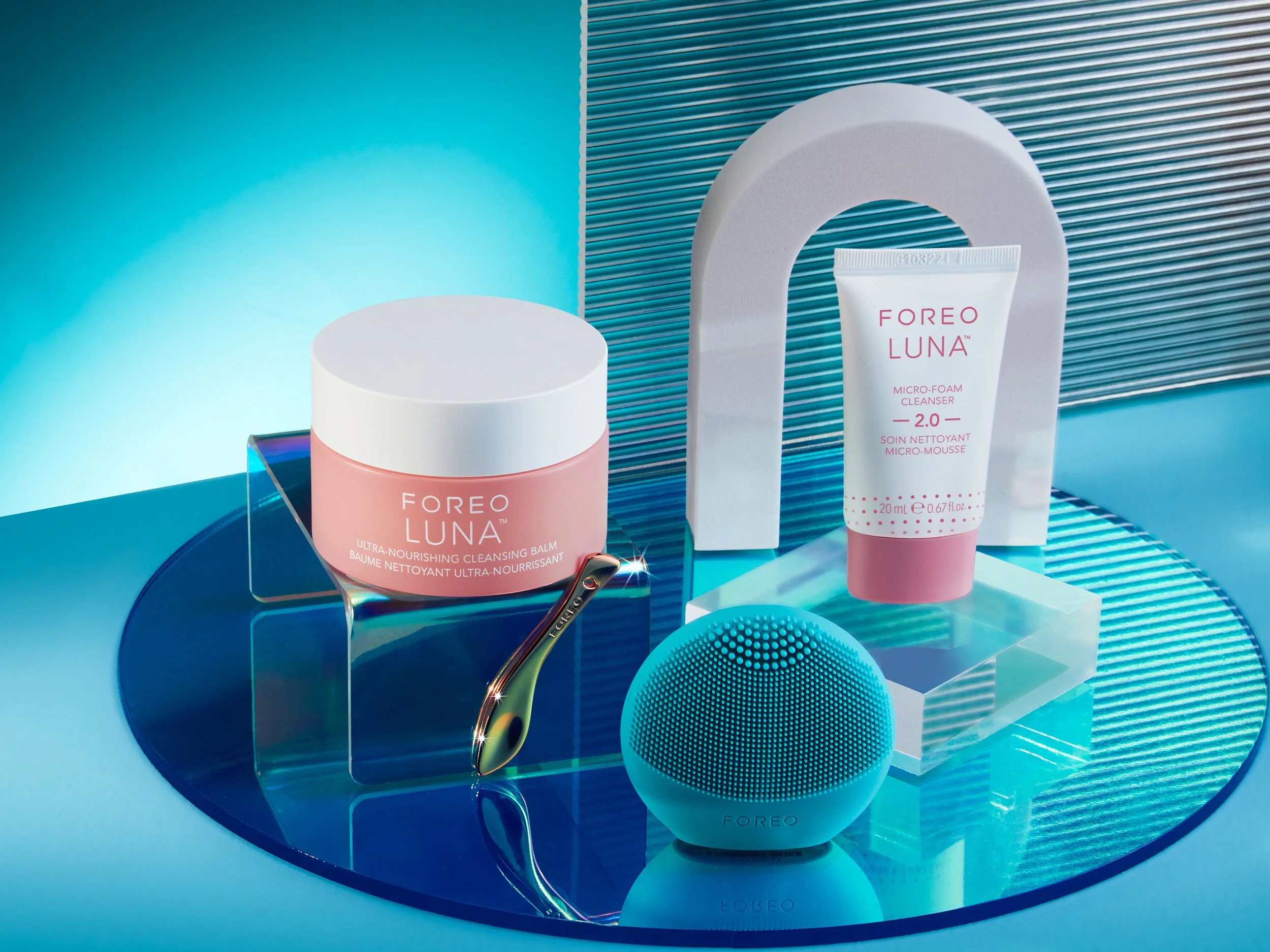 Display of Foreo Luna skincare devices and products on a reflective blue surface with blue and white background. Includes a pink cleansing balm, a micro-foam cleanser, a blue spherical facial cleansing device, and a smaller device, all with sleek, mo