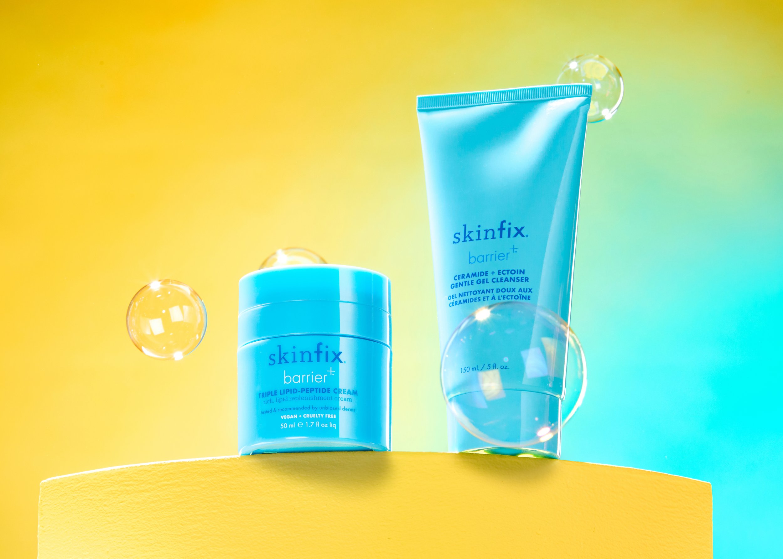 Blue skincare products, a jar and a tube, on a yellow surface with colorful background, with floating bubbles.  premium product photography