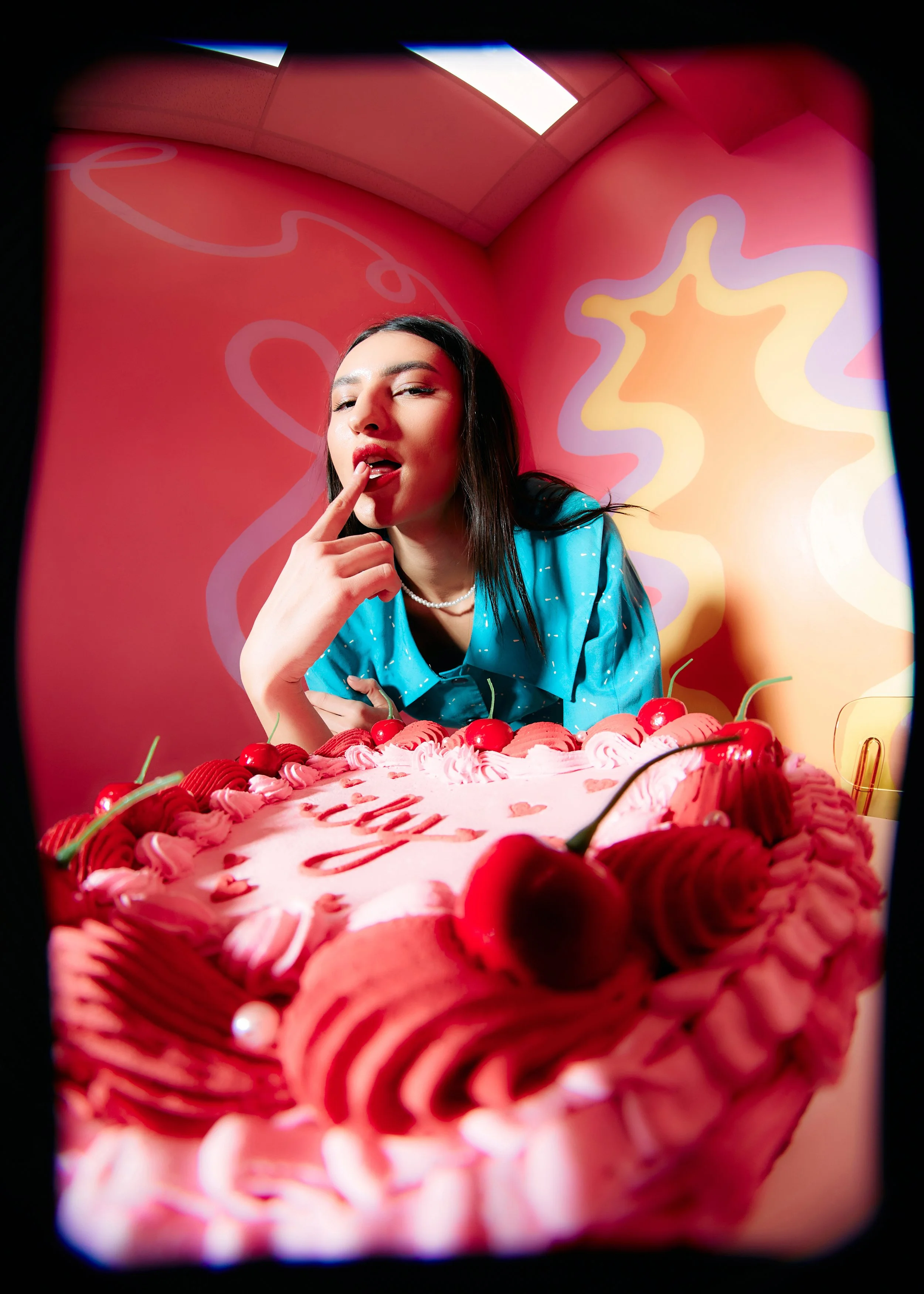 A woman with dark hair, wearing a blue shirt and pearl necklace, posing with a pink birthday cake decorated with cherries, whipped cream, and strawberries. She is touching her lips with her finger in a playful manner, against a vibrant pink, orange, 