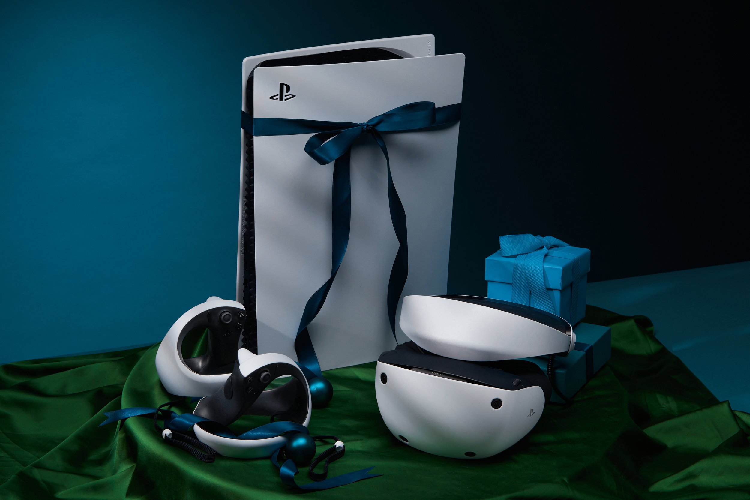 A PlayStation 5 console with a black and white design, wrapped with a blue ribbon, and several virtual reality accessories including a headset and controllers, all placed on a green cloth with a dark background.