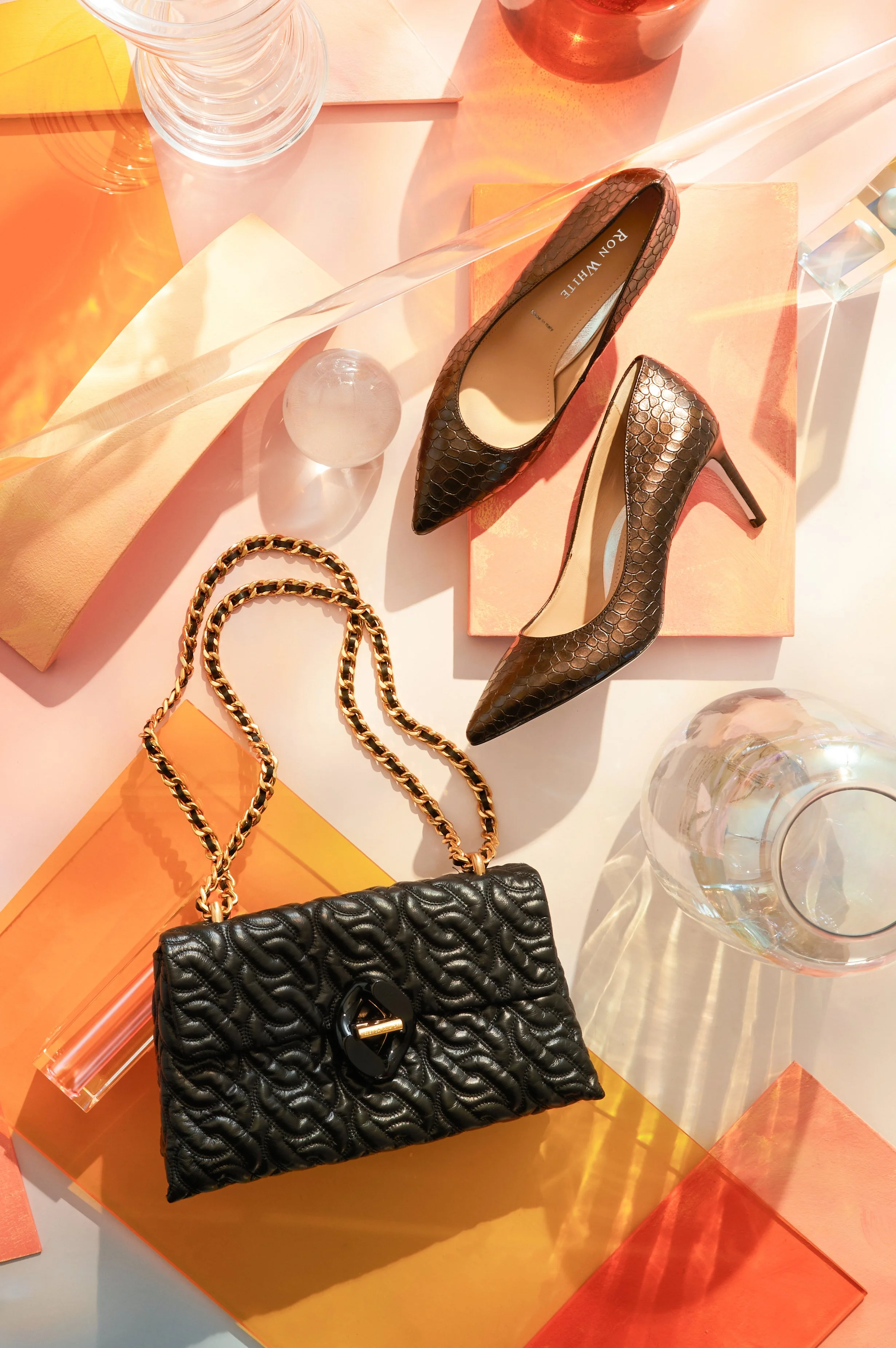 A pair of brown snakeskin high heel shoes, a black quilted handbag with a gold chain strap, a clear glass, and a clear glass sphere on a pastel-colored surface with orange and yellow accents. The shoes are placed on a pink box, and there are addition