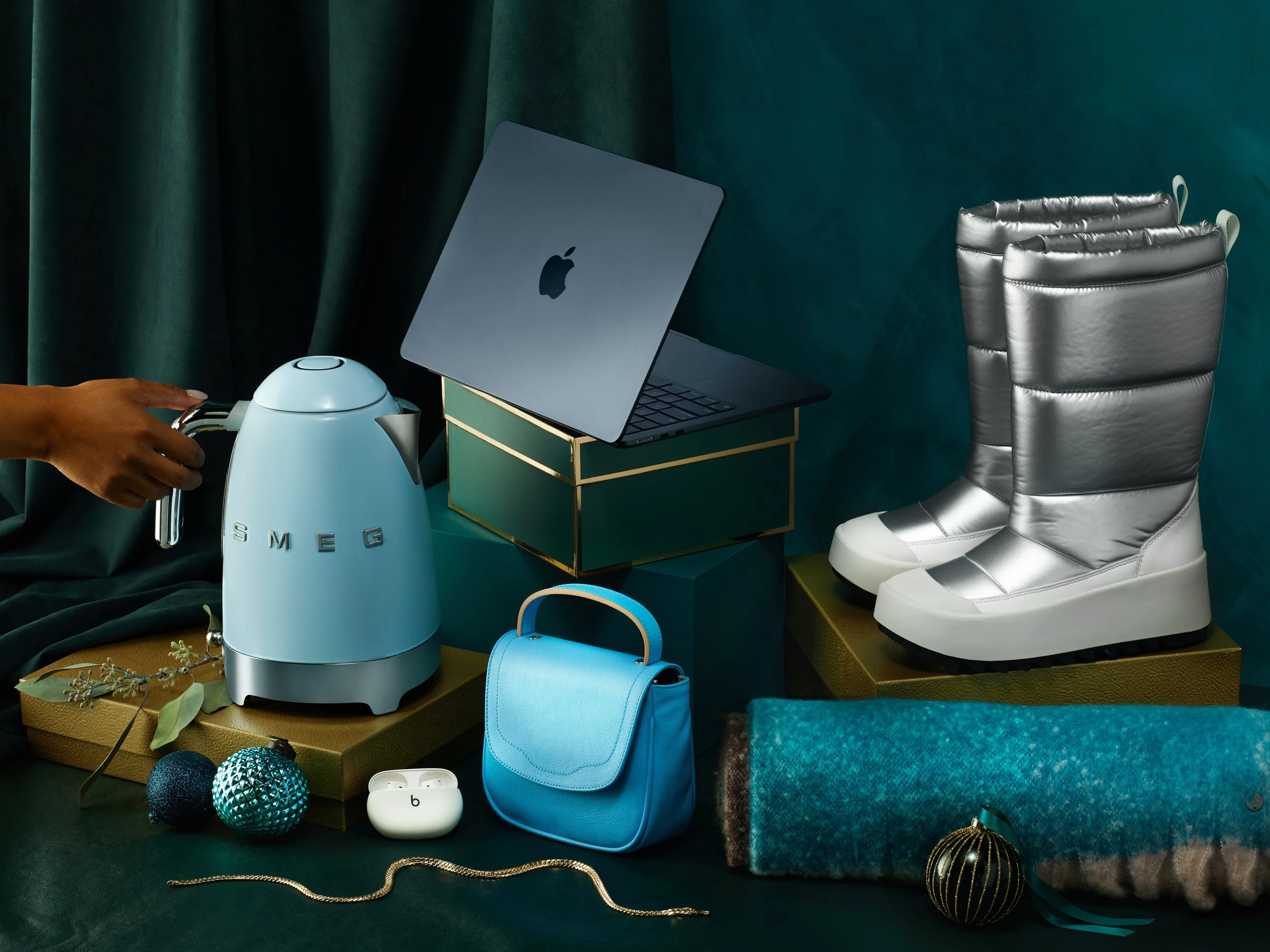 The image features a collection of modern fashion and lifestyle items, including metallic silver winter boots, a pastel blue kettle with 'SMEG' branding, a light blue handbag, a pair of white wireless earbuds, a green box, a laptop, and decorative Ch
