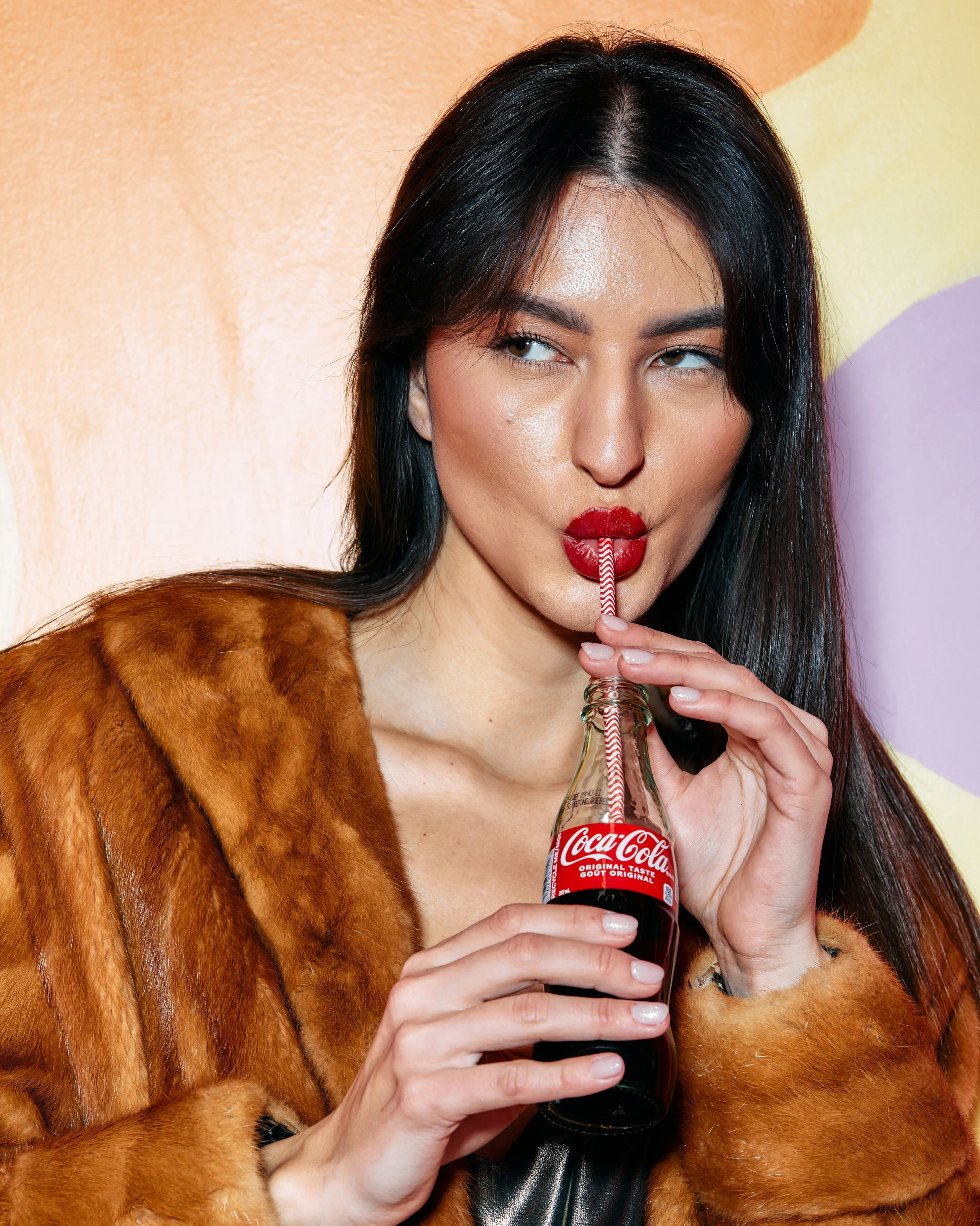 A woman with dark hair and red lipstick drinking from a small Coca-Cola glass bottle using a striped straw, wearing a brown fur coat.