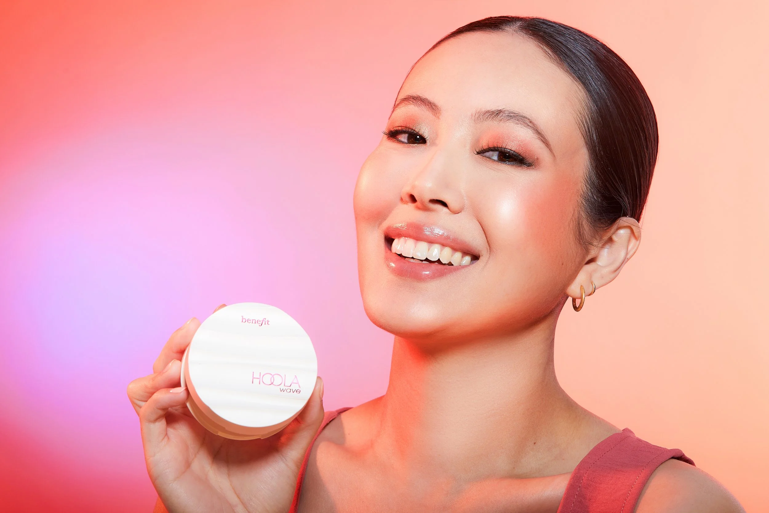 A woman with short dark hair and gold hoop earrings smiling while holding a round container of benefit Hoola Wave bronzer against a pink and peach background.