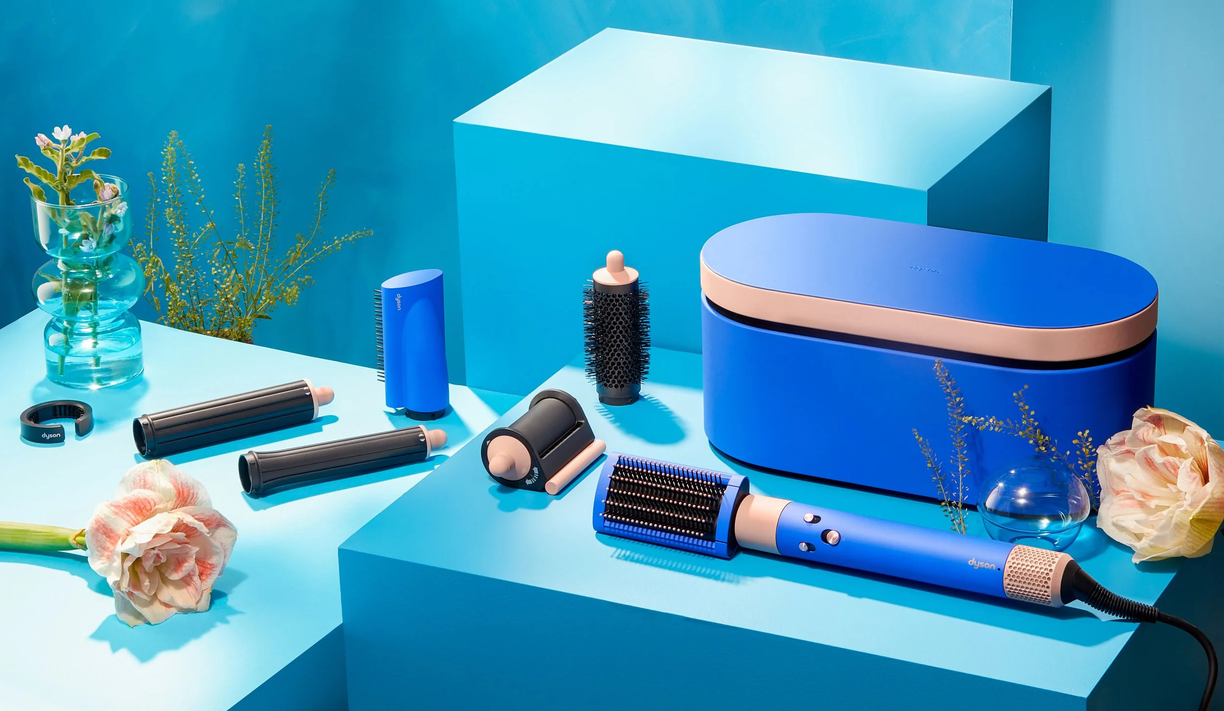 Collection of blue and black Dyson hair styling tools, including a hair dryer, brush, and various attachments, set on a blue background with flowers and plants.