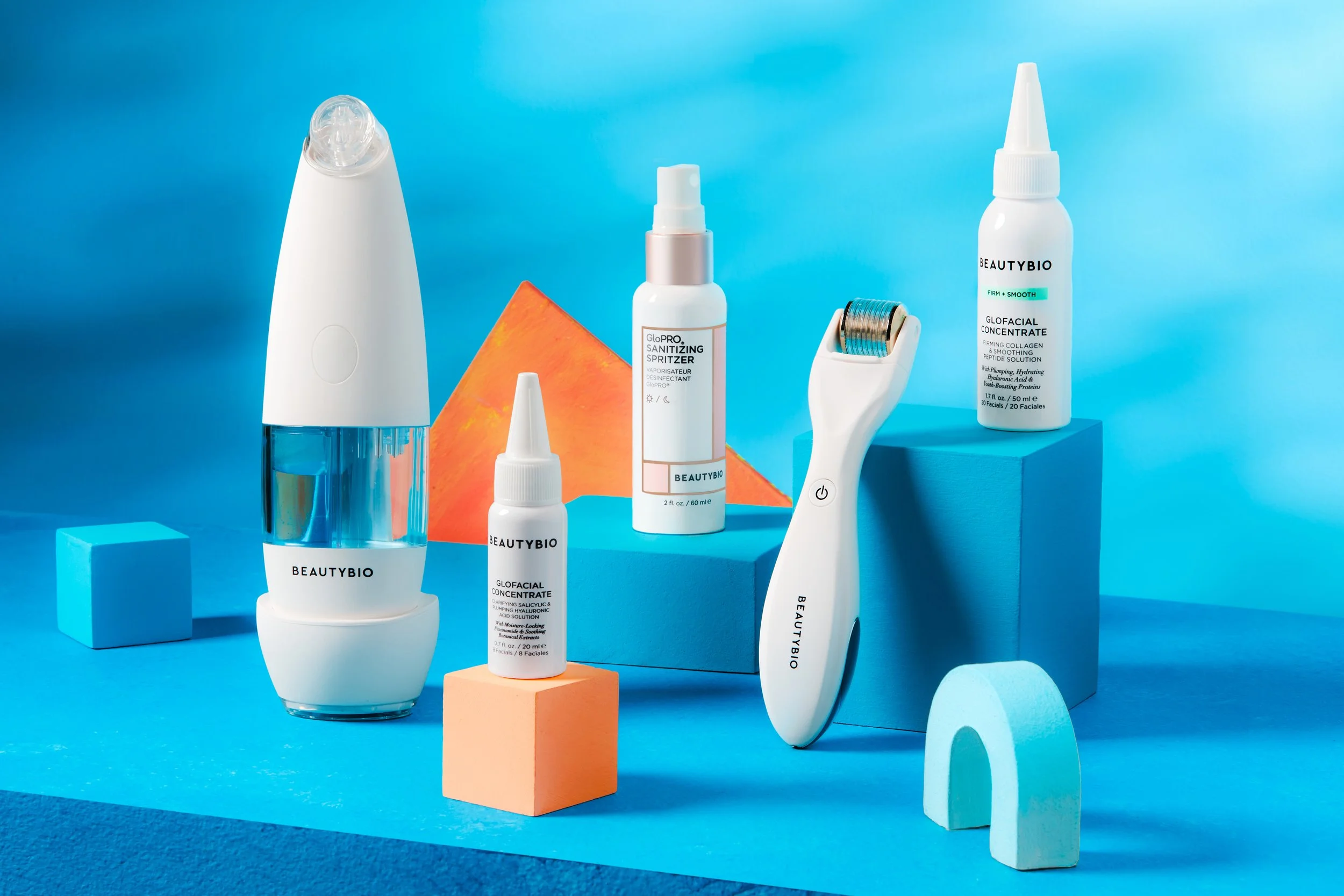 Collection of skincare devices and products on blue background, including a white facial cleansing brush, a small white dropper bottle of GloFacial Concentrate, a facial mist spray, and a roller tool, with geometric blocks and an orange triangular ba