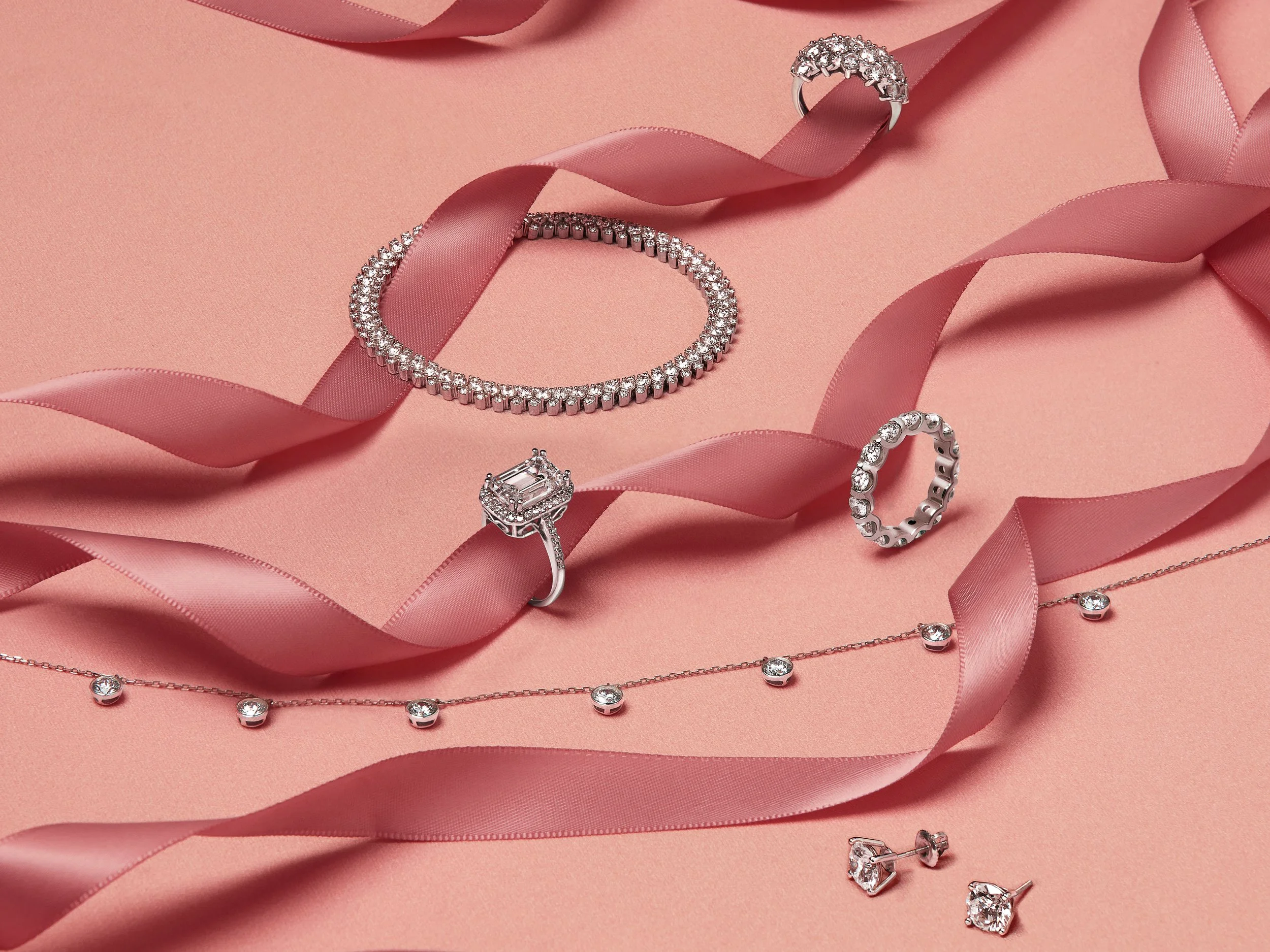 Silver jewelry collection, including rings, necklace, bracelet, and earrings, displayed on pink satin background with pink ribbon.