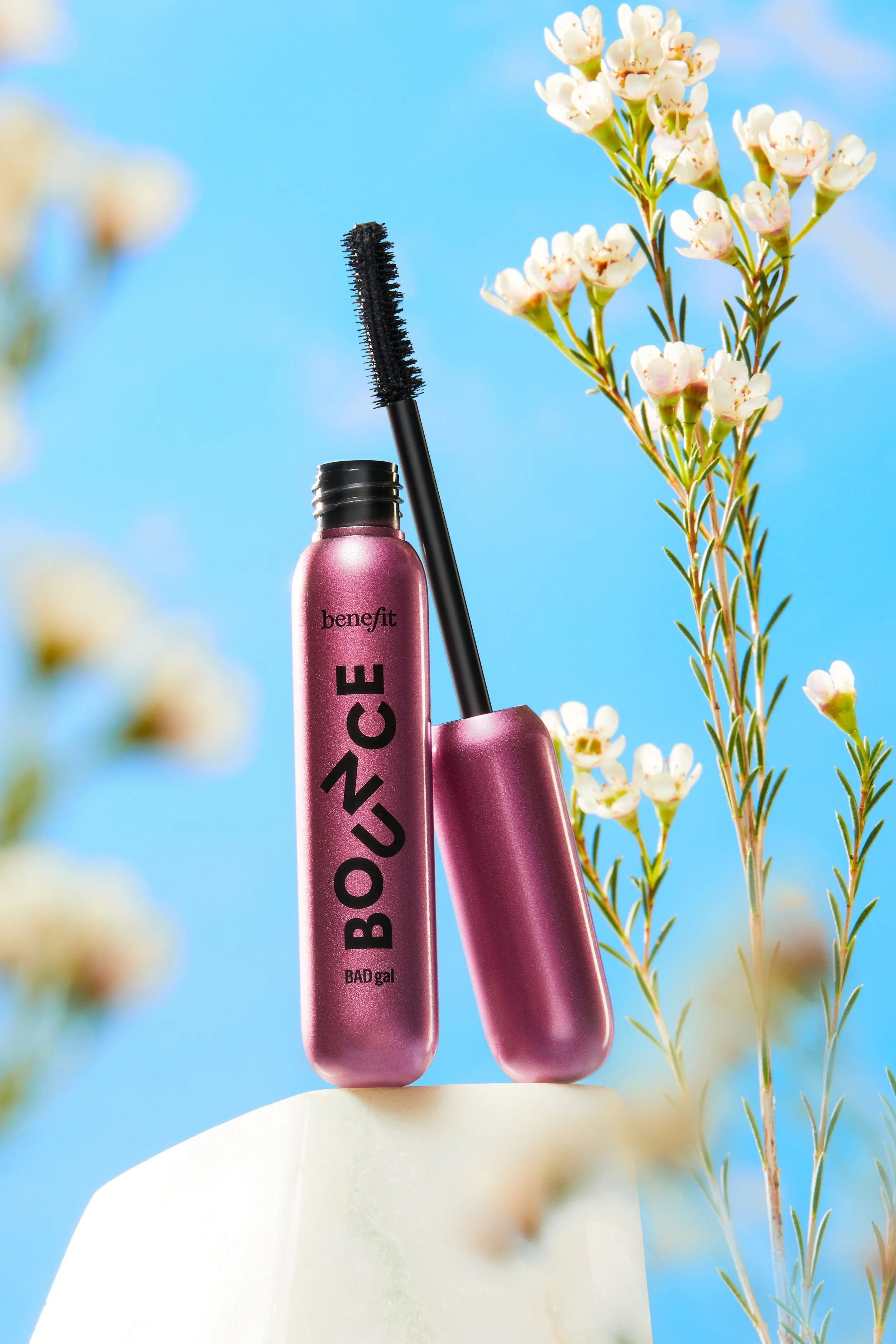 Benefit BADgal BOUCE mascara with wand inserted, set against a blue sky background, with white flowers and green foliage around.