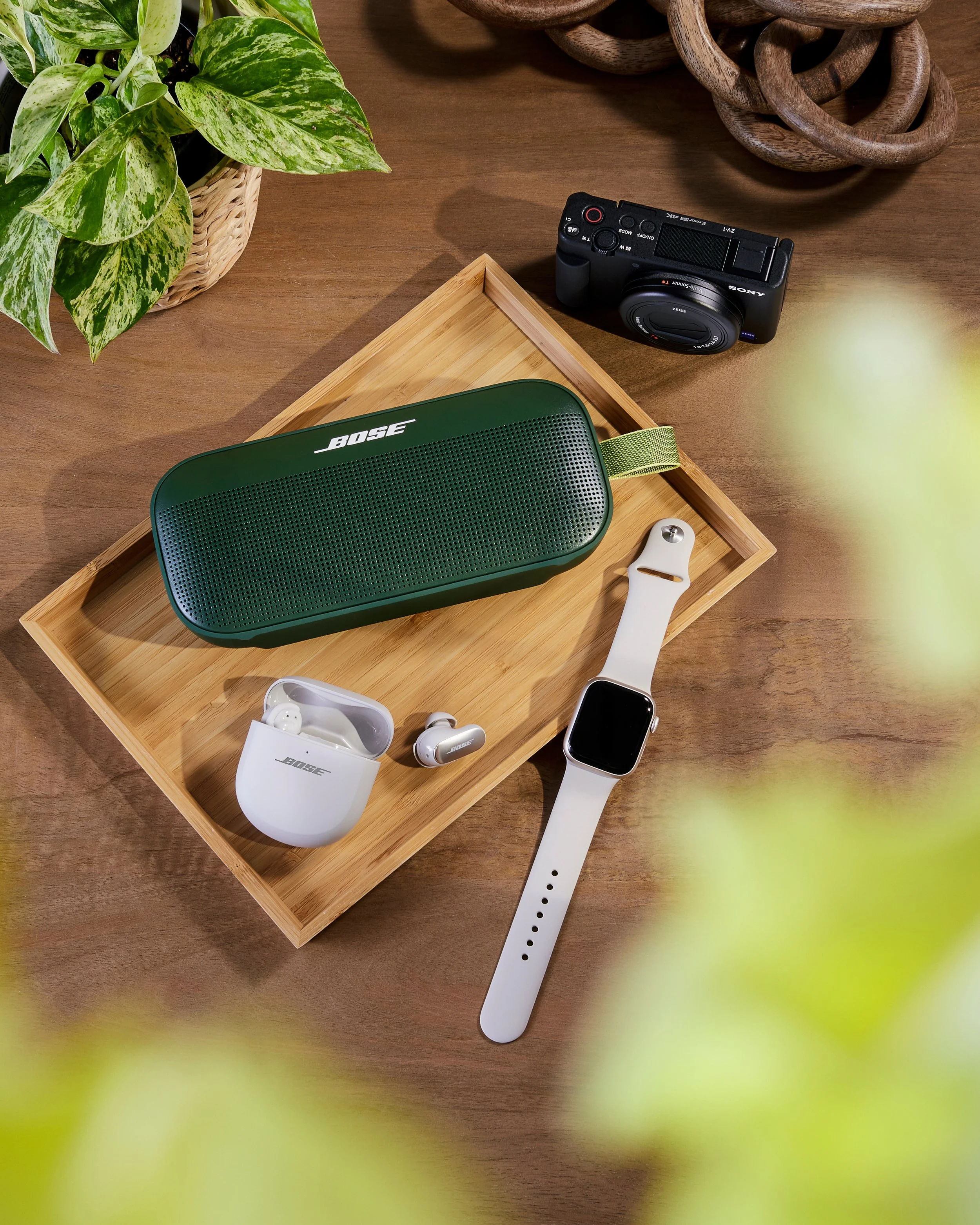 A flat lay of electronic gadgets on a wooden table includes a green Bose portable speaker, a pair of white Bose wireless earbuds in a charging case, a white smartwatch, and a black digital camera. There is also a wooden tray, a potted plant with vari