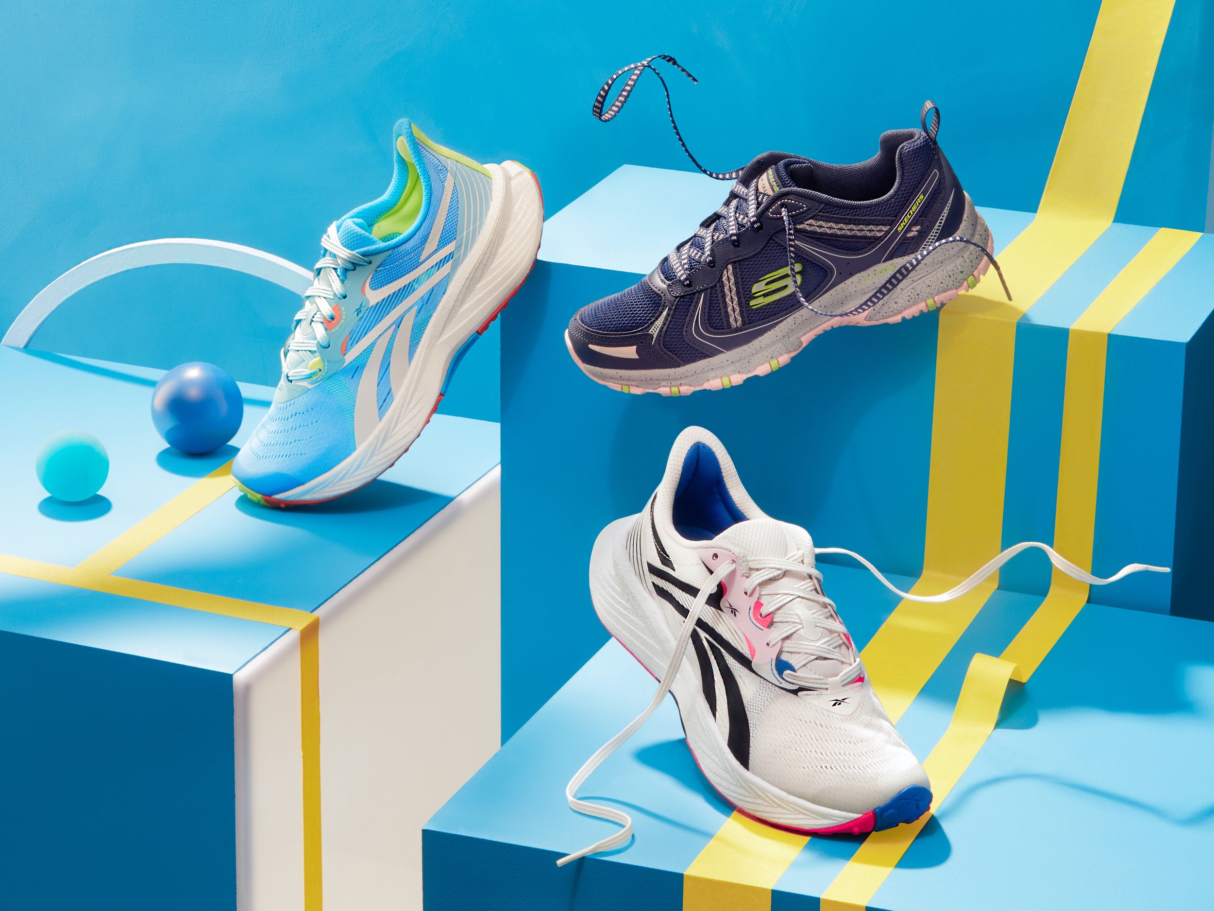 Three athletic running shoes displayed on colorful blue and yellow geometric platforms with decorative spheres and a curved white arch in the background.
