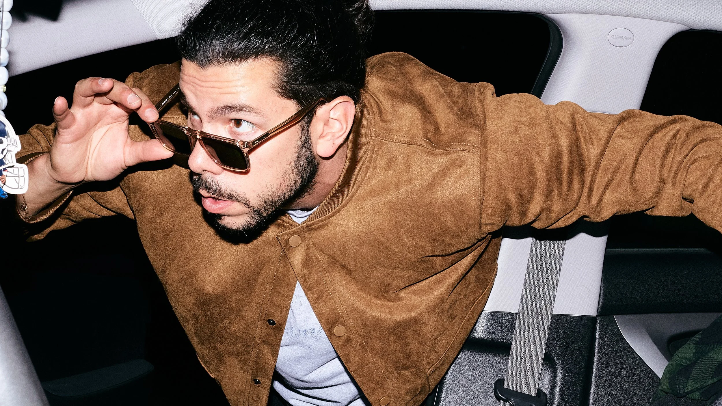 A man with dark hair and a beard adjusting his sunglasses while sitting in the back seat of a car.