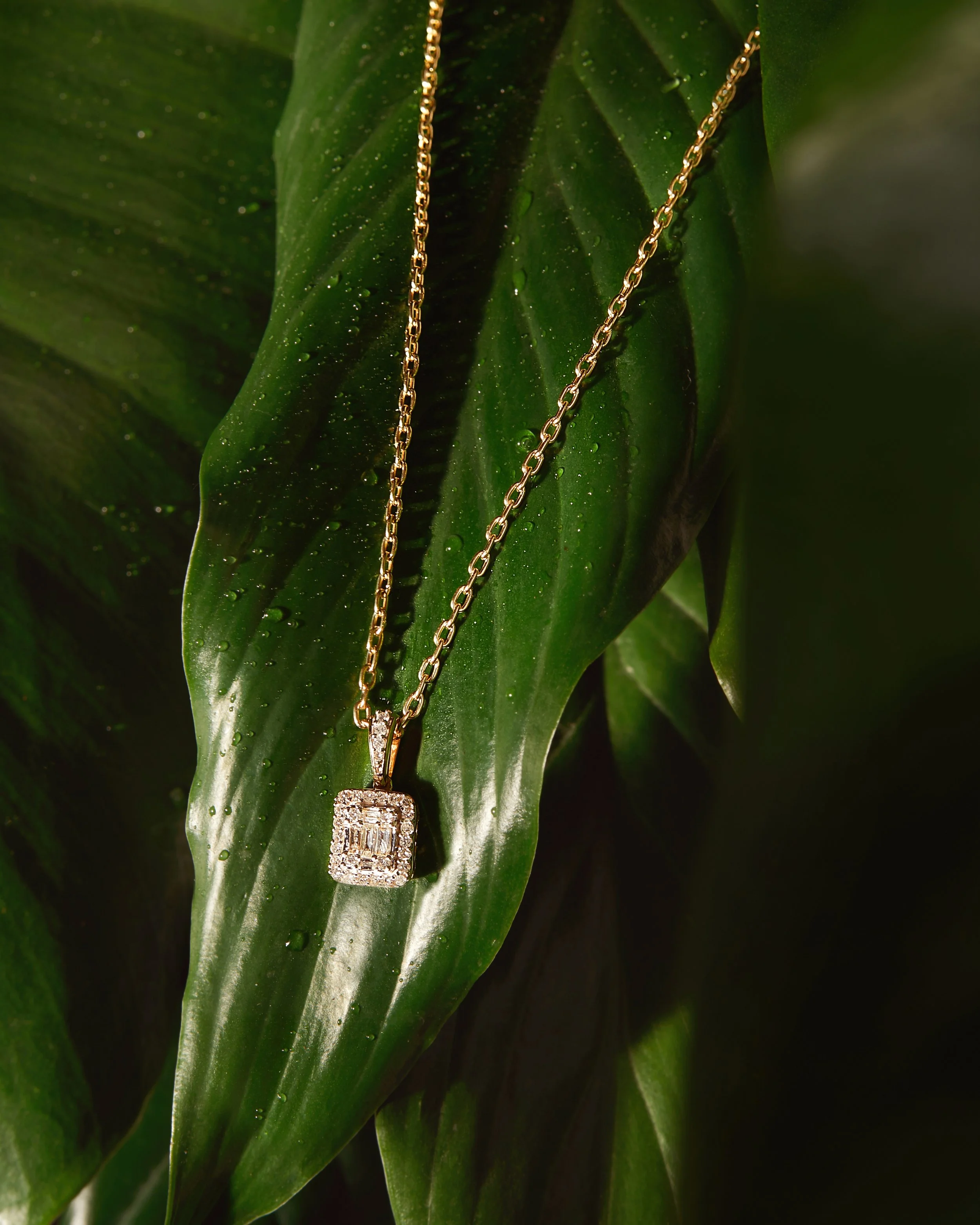Gold chain necklace with a square pendant featuring diamonds, hanging on a wet green leaf.
