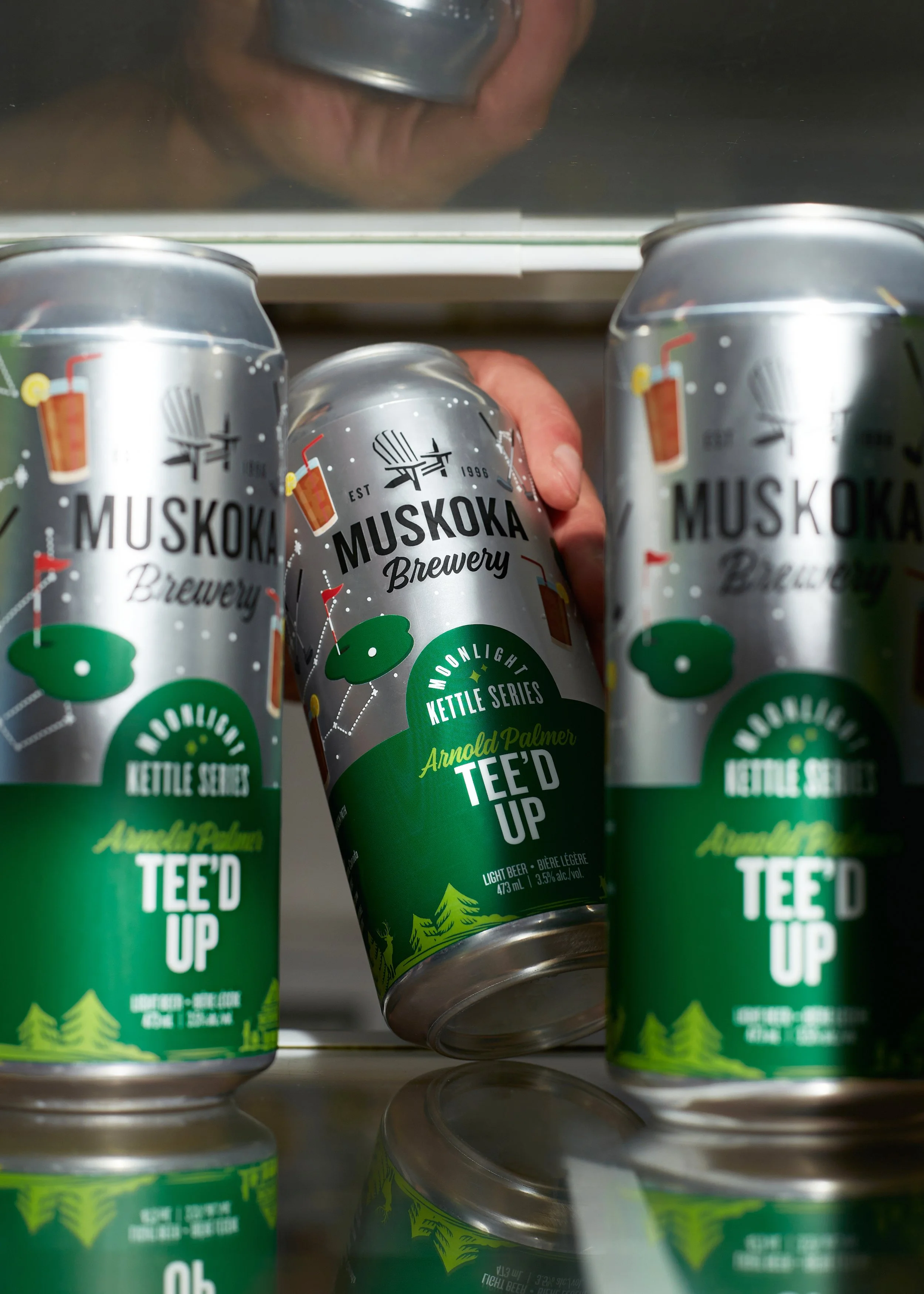 Three cans of Muskoka Brewery's Tee'D Up beer on a refrigerator shelf, with one can being held by a hand, viewed from below. Lifestyle beverage shot