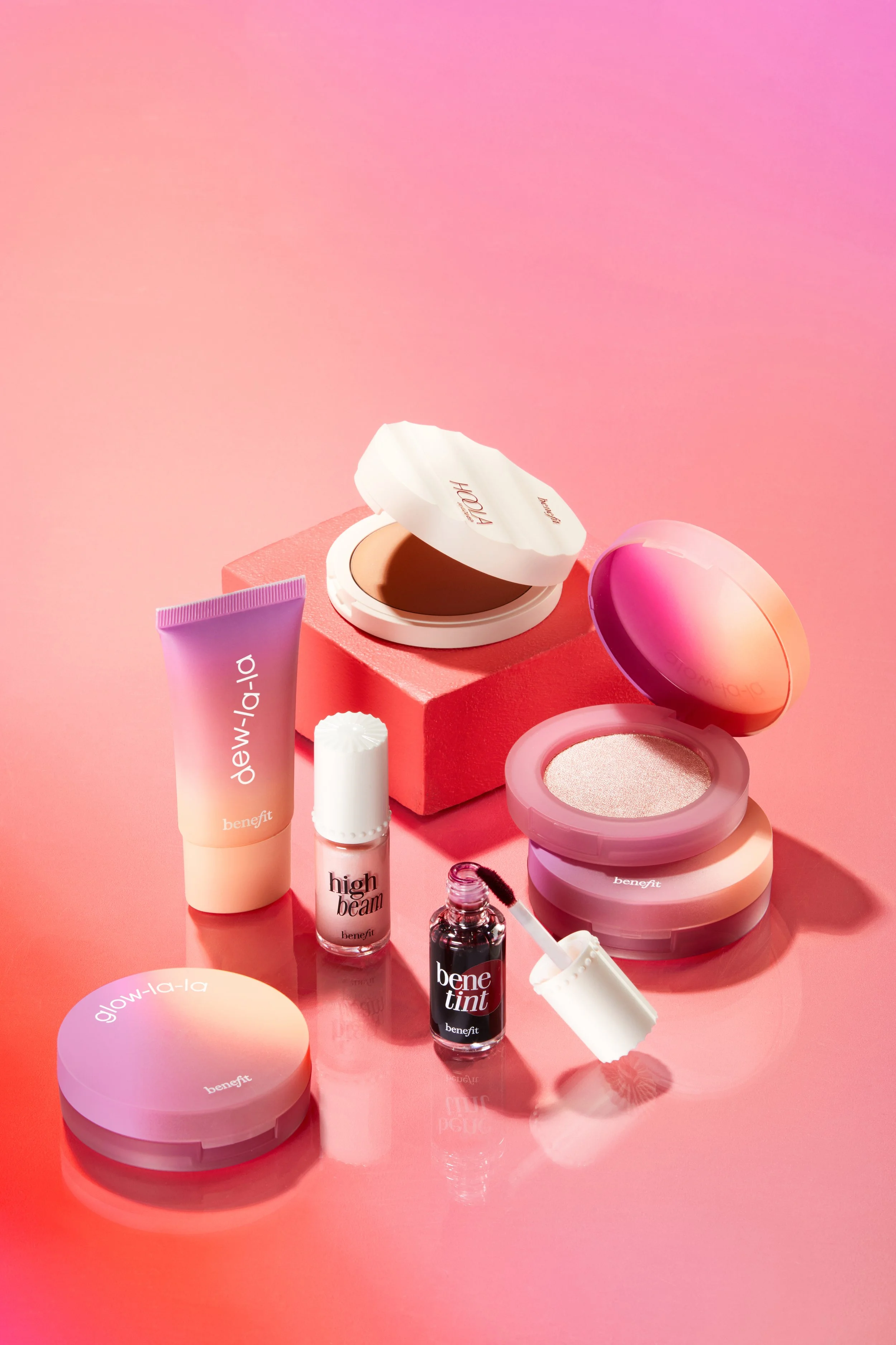 Collection of Benefit cosmetics products on pink background, including blush, primer, concealer, lip tint, and powder.