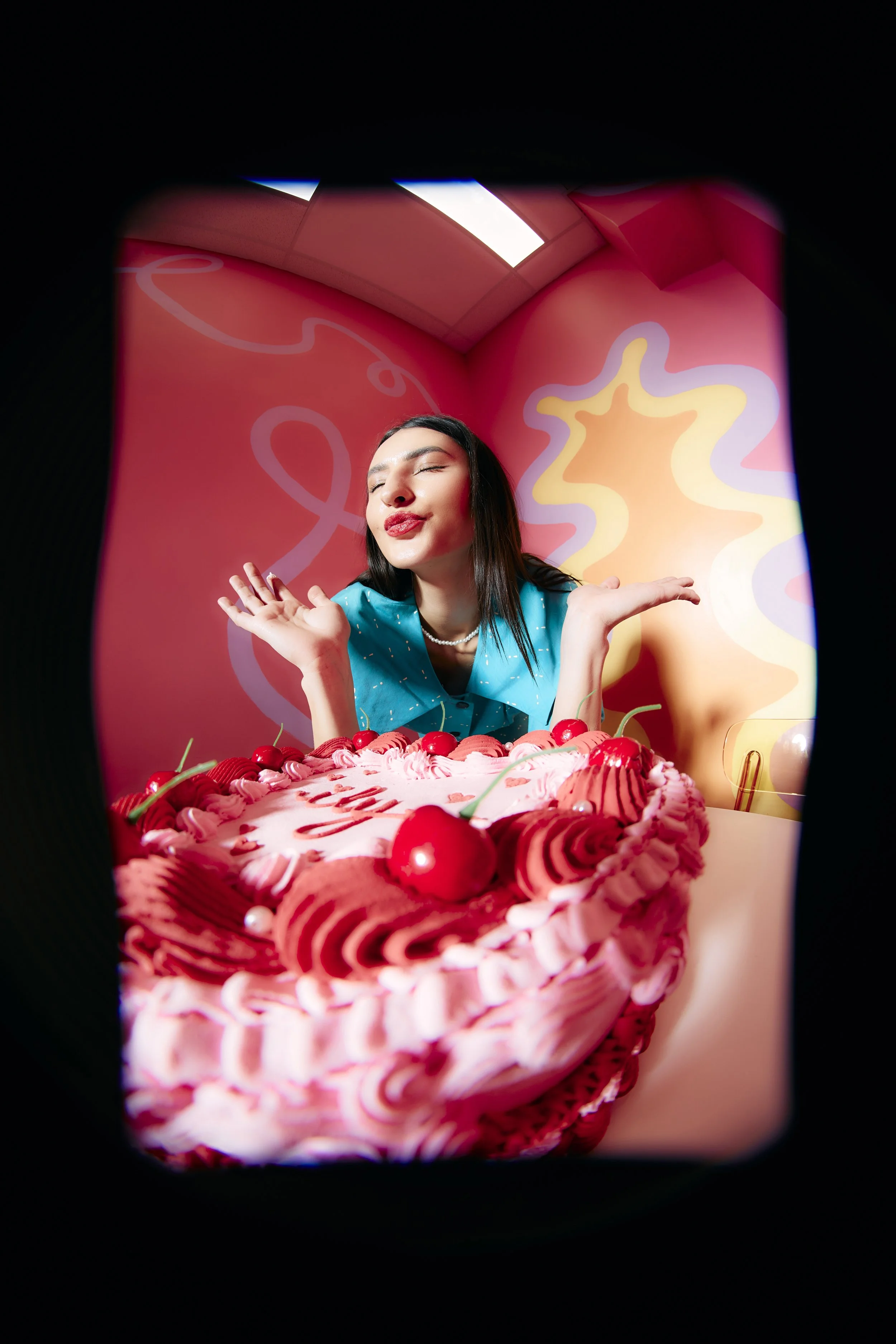 A young woman with dark hair and red lipstick makes a kissy face with eyes closed, at a celebration with a large pink cake decorated with cherries and pink frosting. She wears a blue top with white polka dots and a pearl necklace, in front of a pink 