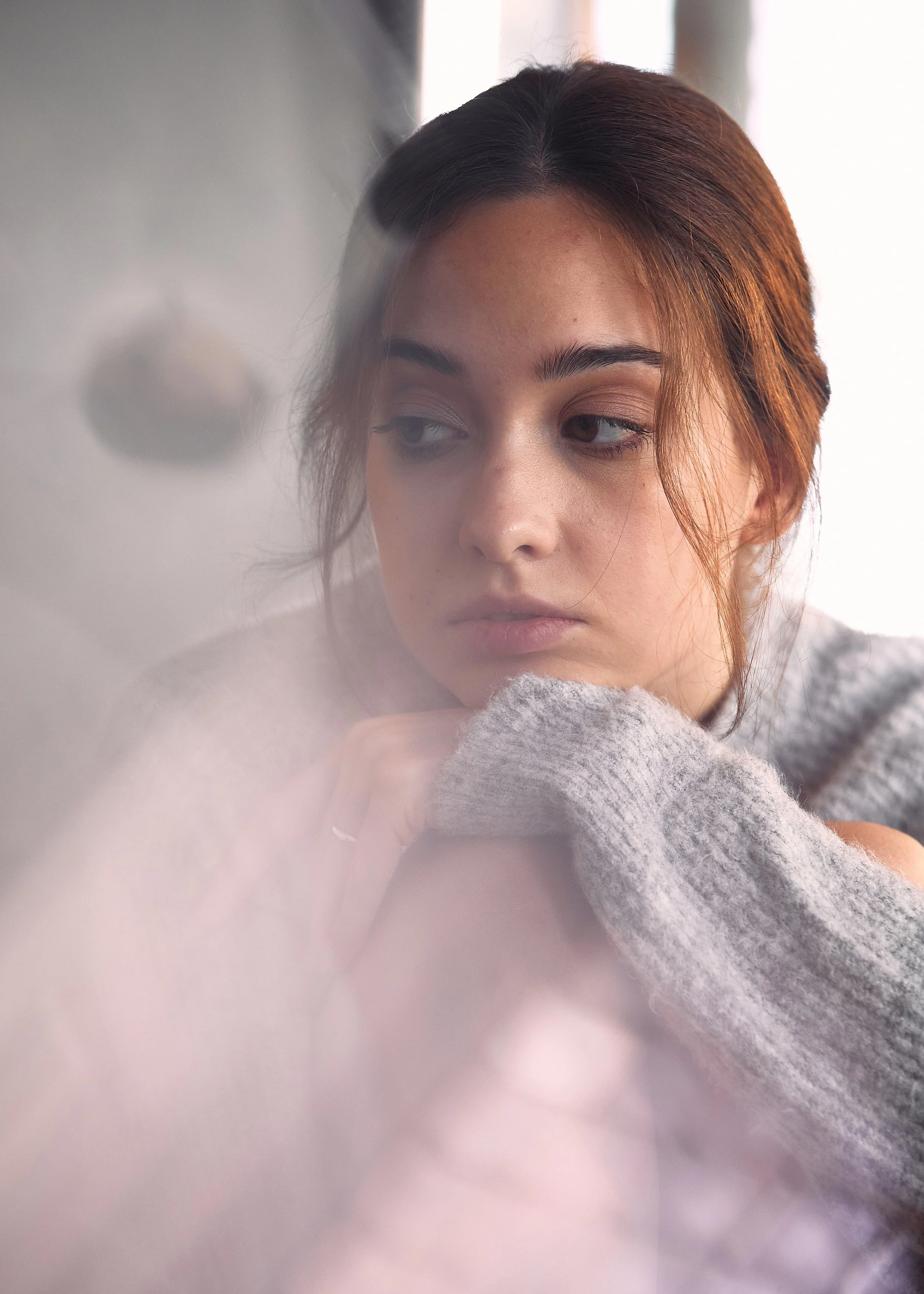 A young woman with brown hair looking down, resting her chin on her hand, with a contemplative expression, wearing a gray sweater, behind a slightly foggy or frosted glass surface.