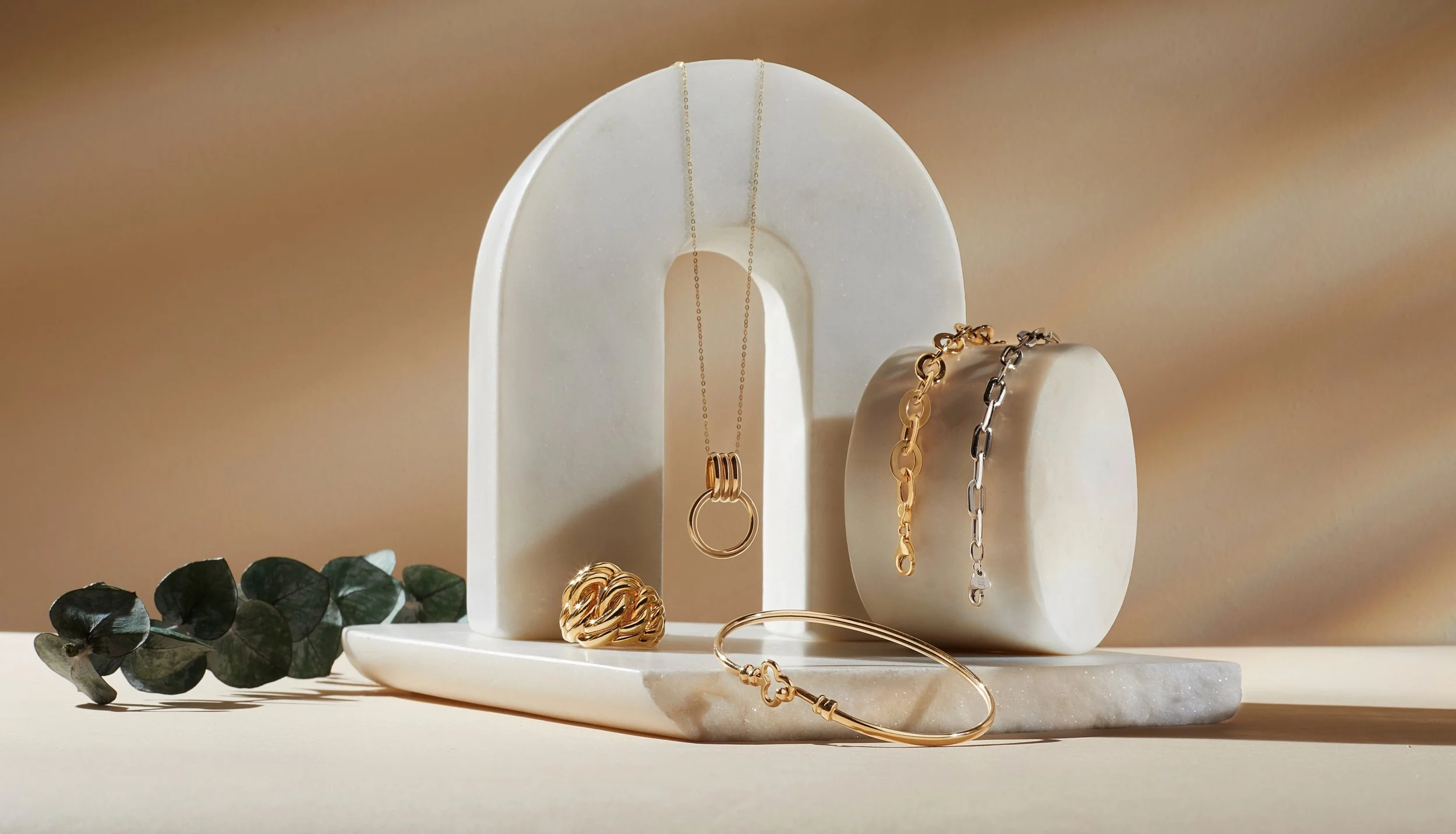 Gold and silver jewelry including chains, rings, and bracelets displayed on white arched and cylindrical stands with a small sprig of eucalyptus on the table.