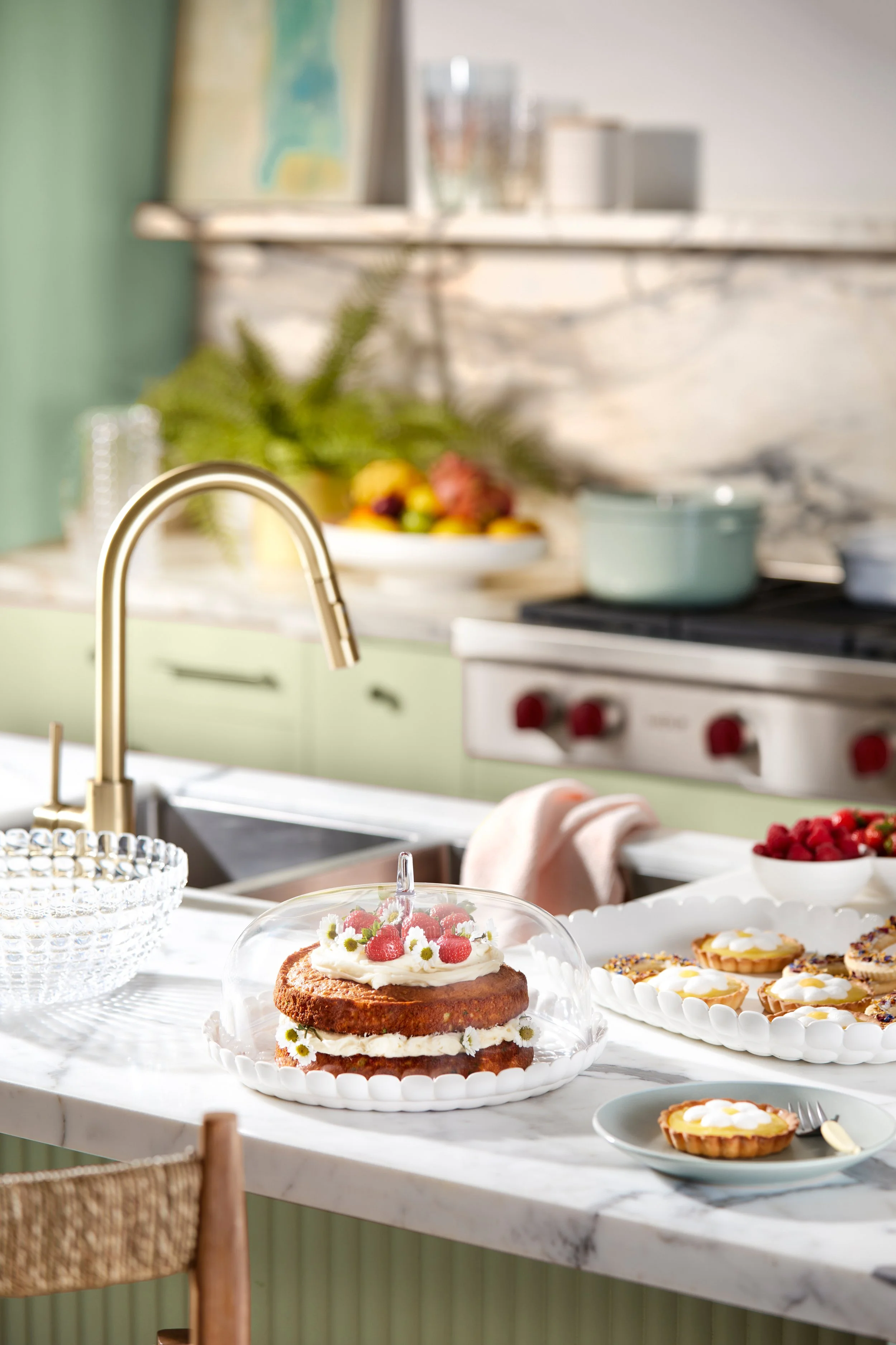 A kitchen counter with a glass cake dome covering a layered cake decorated with berries and flowers, surrounded by plates of tarts and bowls of berries.