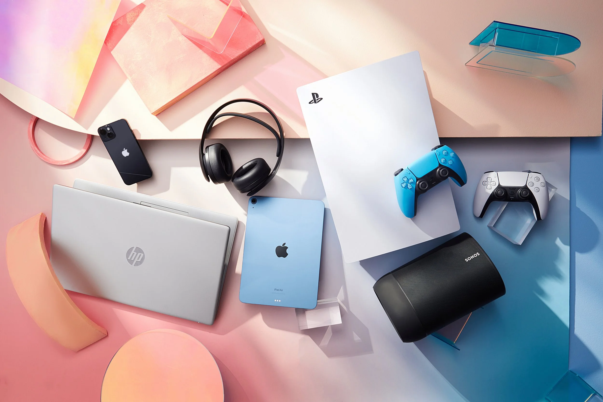 Collection of various electronic devices including a laptop, iPad Air, iPhone, gaming controllers, headphones, and a portable speaker arranged on colorful paper backgrounds.