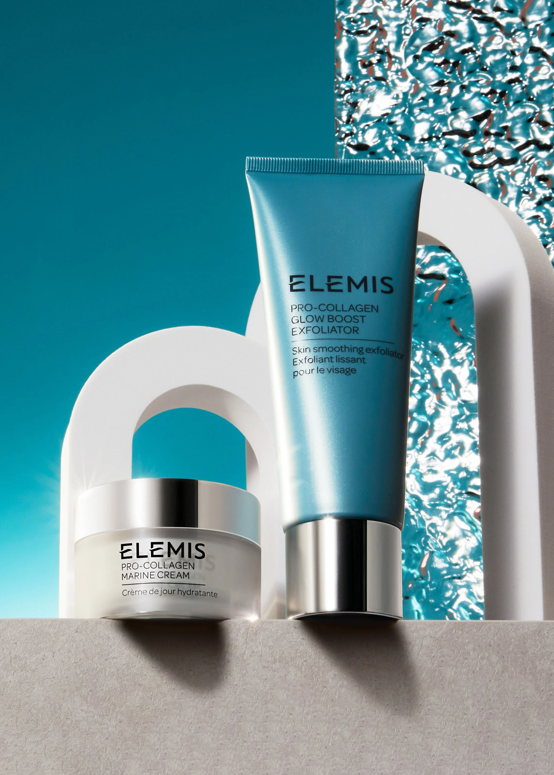 Two Elemis skincare products, a marine cream jar and a collagen glow boost exfoliator tube, displayed on a concrete surface with a blue and white abstract background and white decorative arches.
