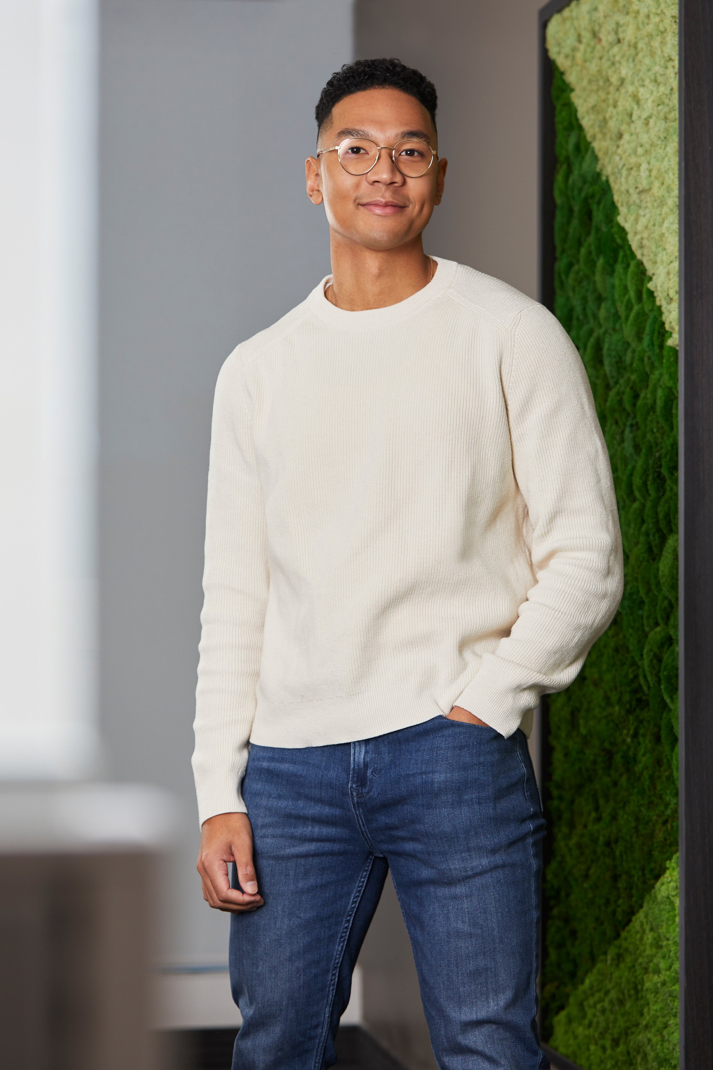 A young man with short curly black hair and glasses, wearing a cream-colored sweater and blue jeans, standing indoors near a green moss wall with a confident expression. Professional branding photography