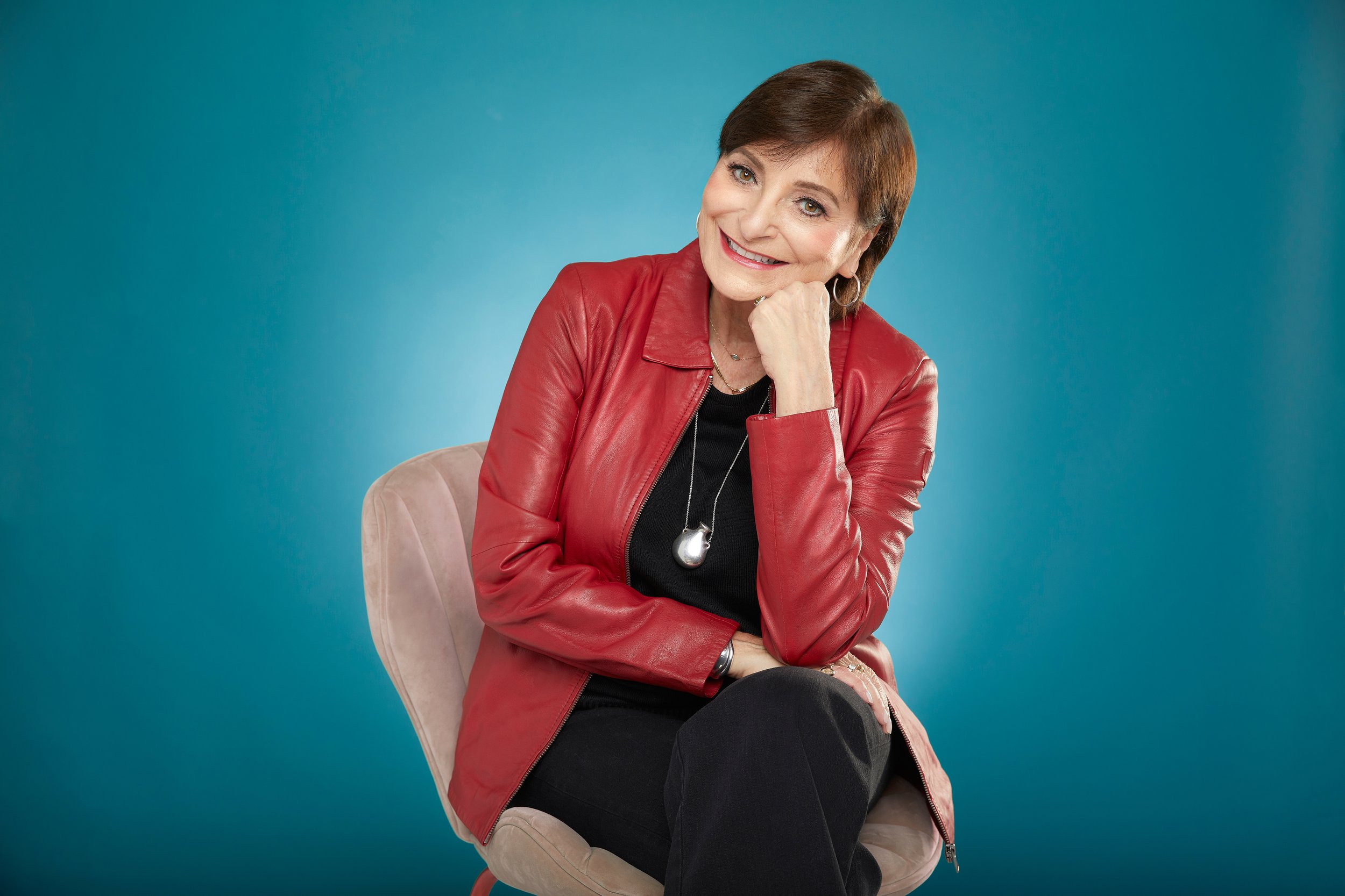 Woman with short brown hair wearing a red leather jacket and black top, sitting on a beige chair against a teal background, smiling and resting her chin on her hand.