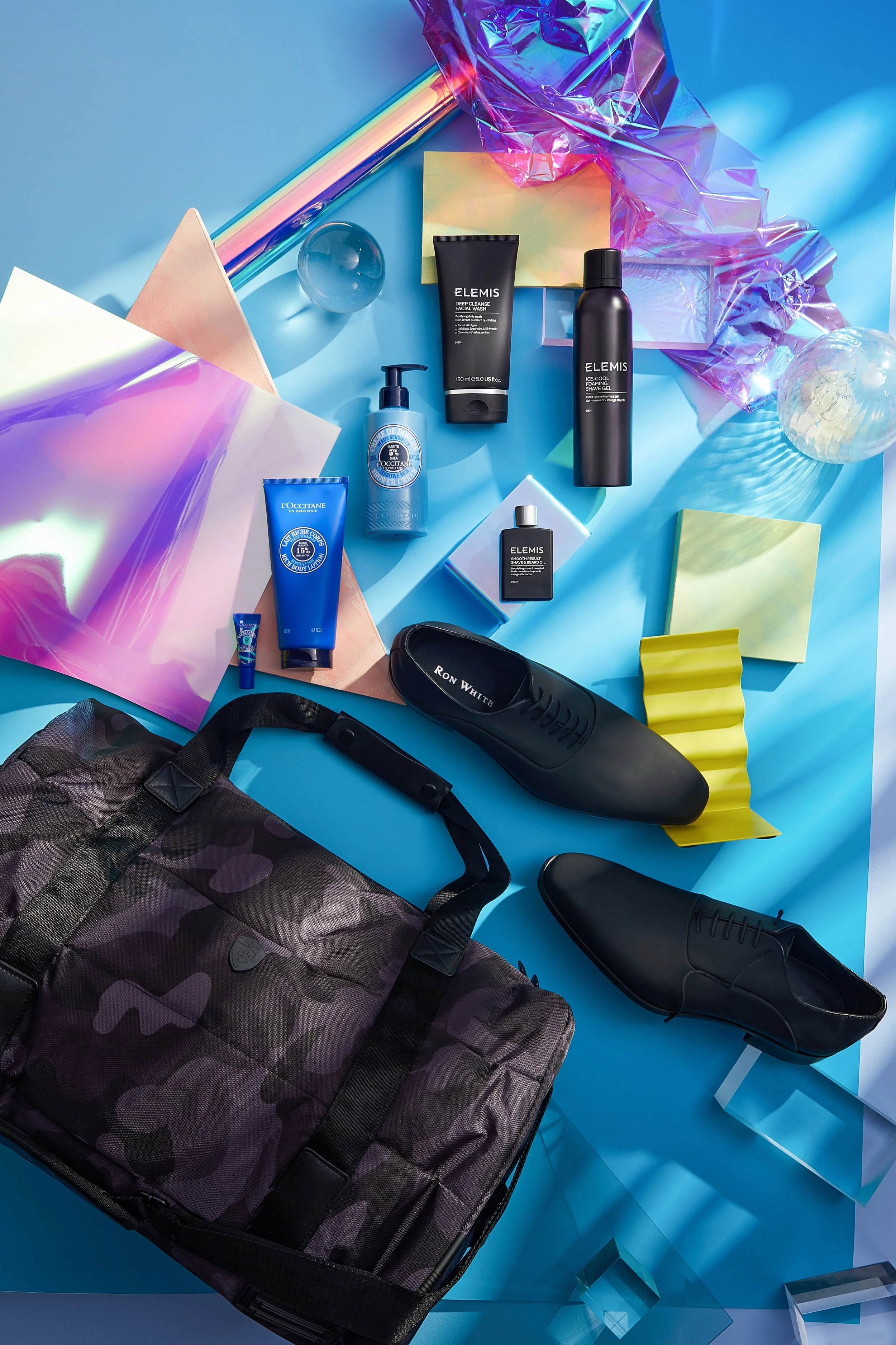 Fashion and grooming items arranged on a blue surface, including a black Ron White shoes, a camo pattern bag, skincare and grooming products, colorful paper, and decorative objects.