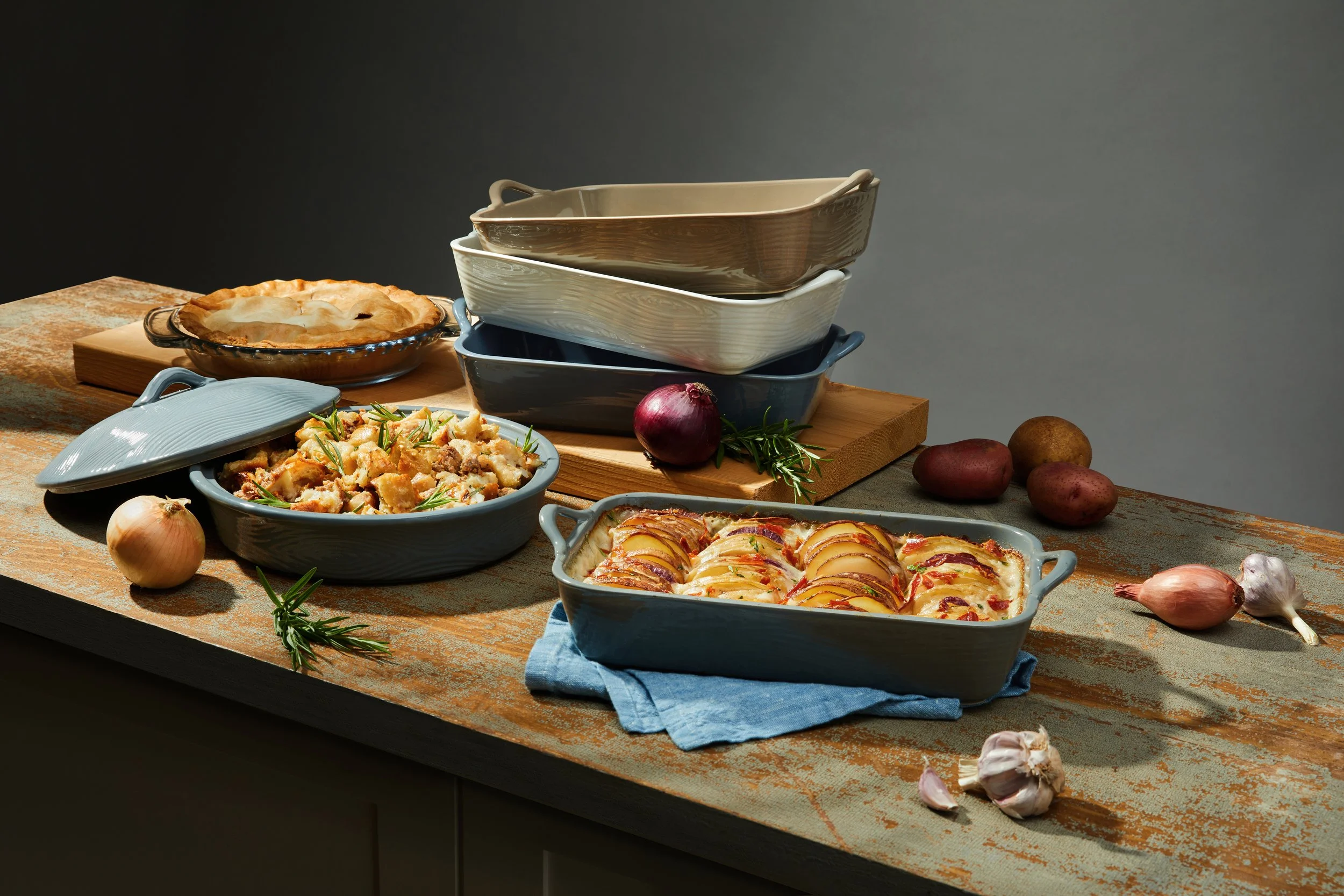 A rustic kitchen table with various baked dishes in ceramic and porcelain bowls, including a potato and stuffing casserole, an apple crisp, and a savory pie, surrounded by onions, garlic, and herbs.