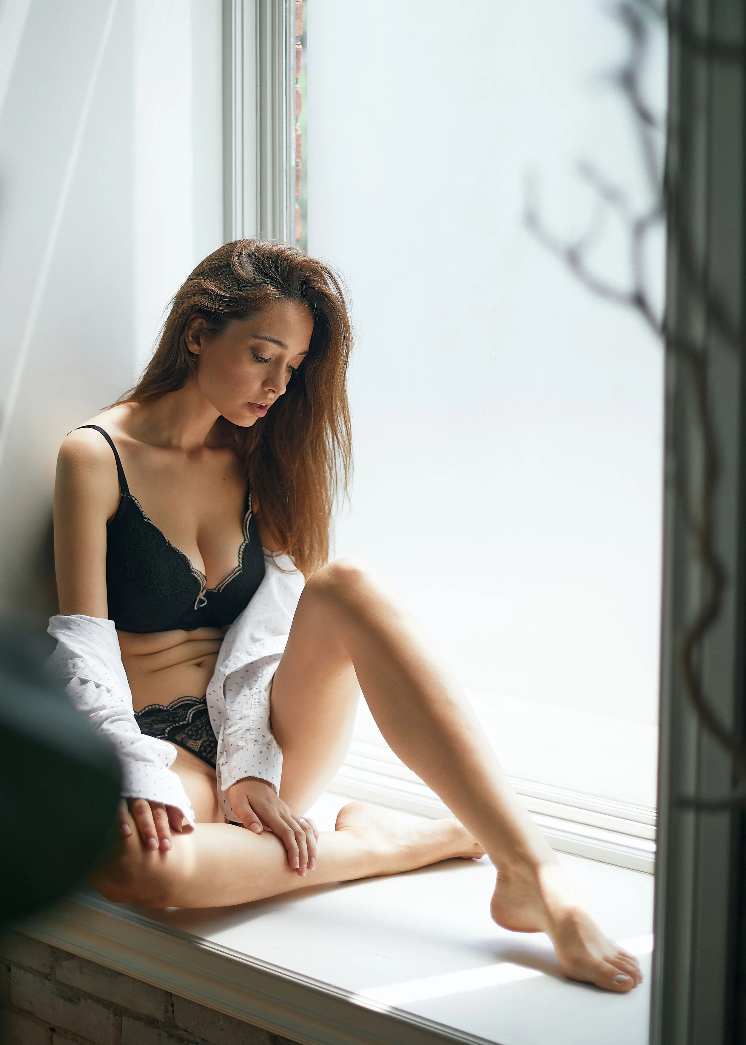 A woman with long brown hair sitting by a window, wearing black lingerie and an open white shirt, looking down with a contemplative expression.