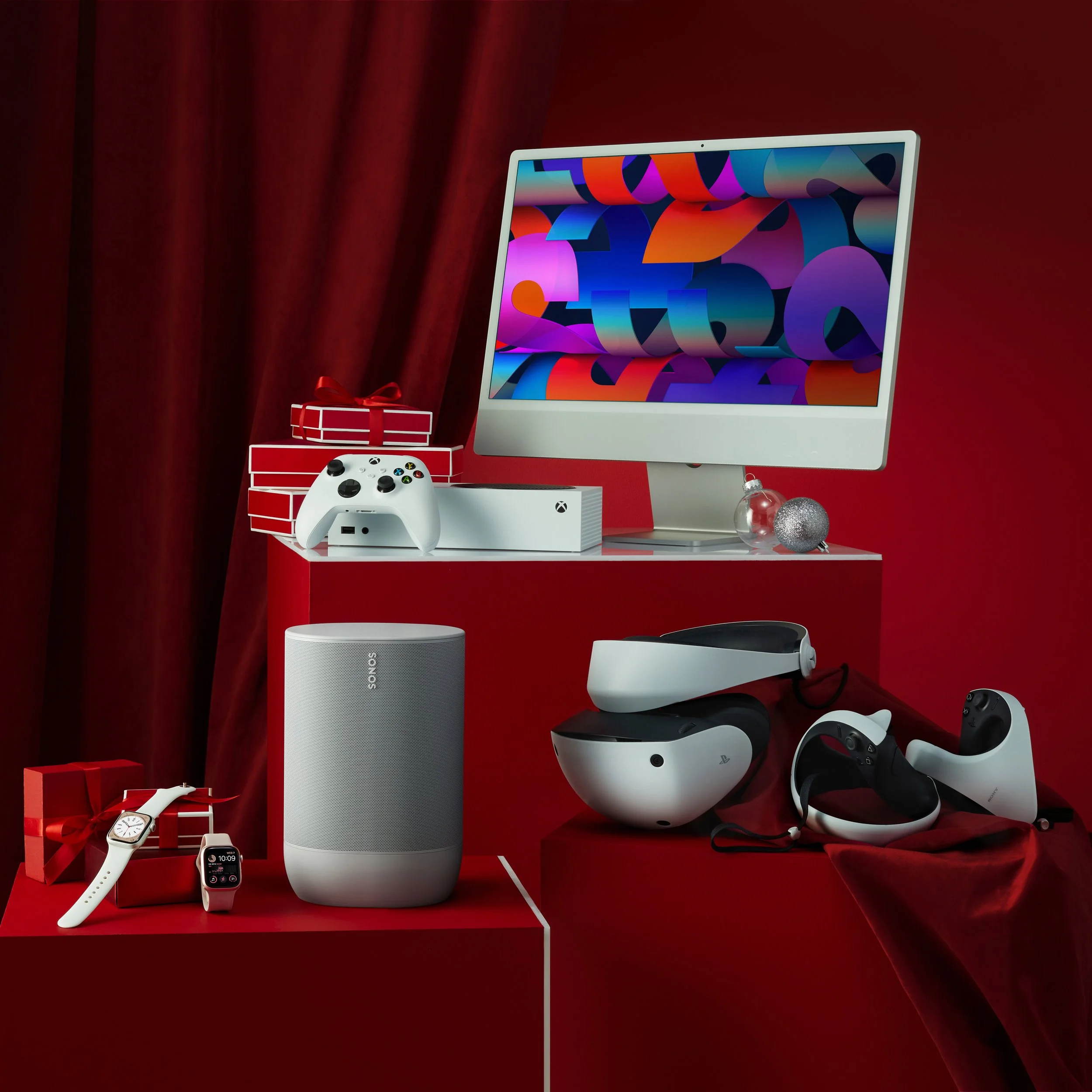 A collection of Apple and Sony electronic devices including a desktop monitor with colorful display, gaming controllers, a smart speaker, a virtual reality headset, and a smartwatch, all set against a red background with Christmas-themed decorations.