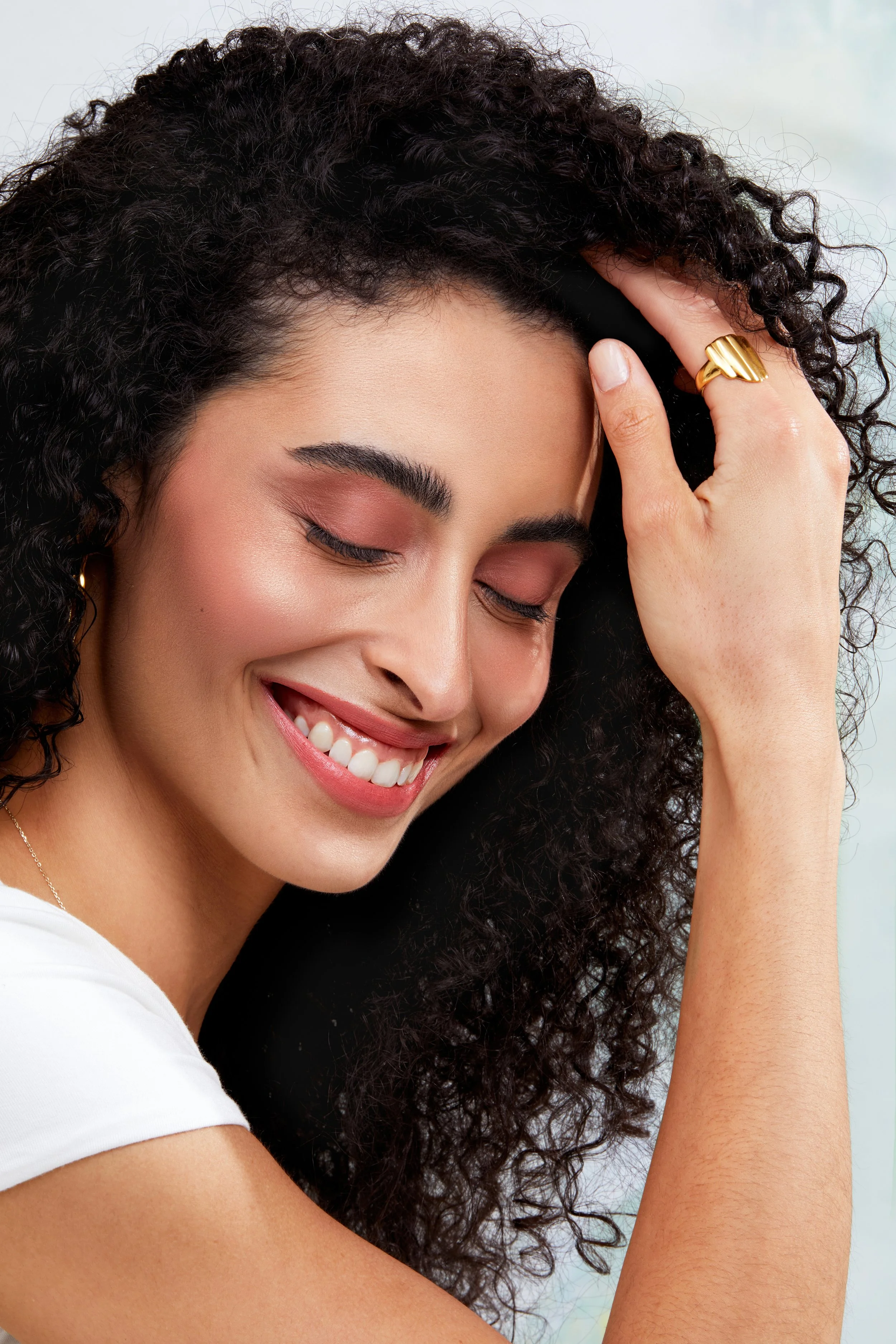 A woman with curly hair smiling with her eyes closed, wearing a white top and gold jewelry.