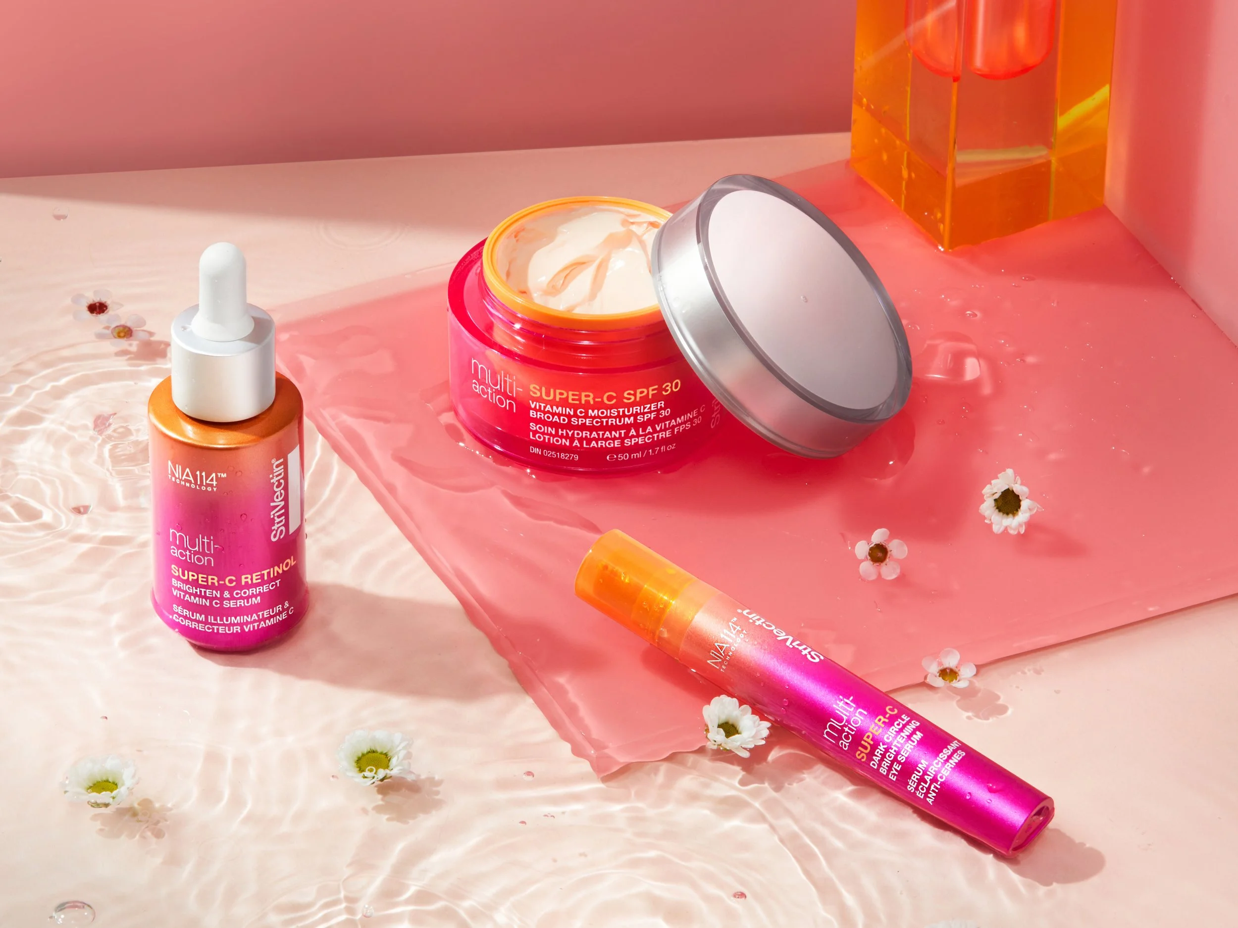 Skincare products including a vitamin C serum, moisturizer with SPF 30, and another serum, arranged on a pink surface with small white daisies and water reflections.