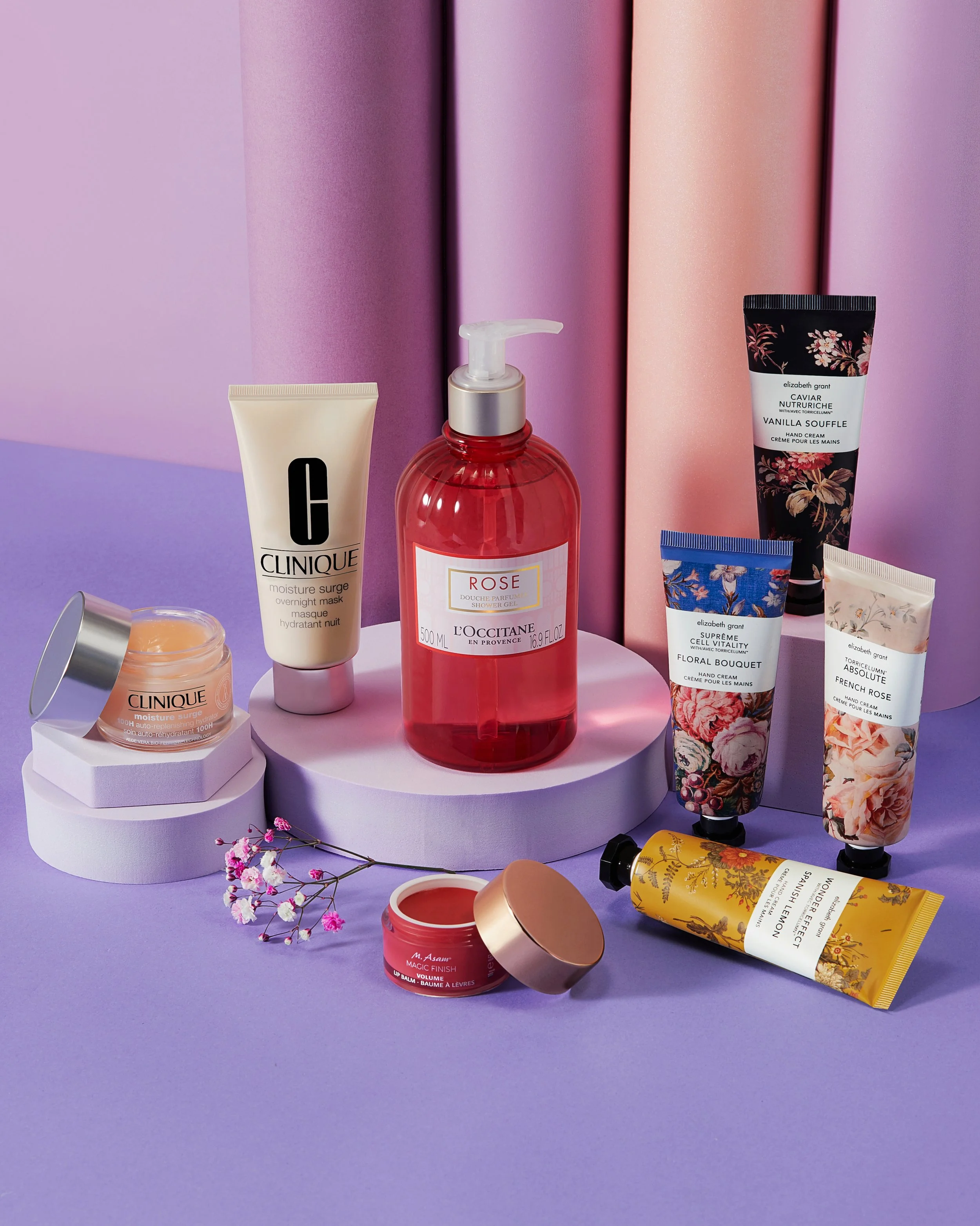 Collection of skincare and cosmetic products including Clinique face mask, L'Occitane rose shower gel, hand creams with floral designs, a lip balm, and a yellow tube, arranged on white platforms with pink and purple background and small pink flowers 