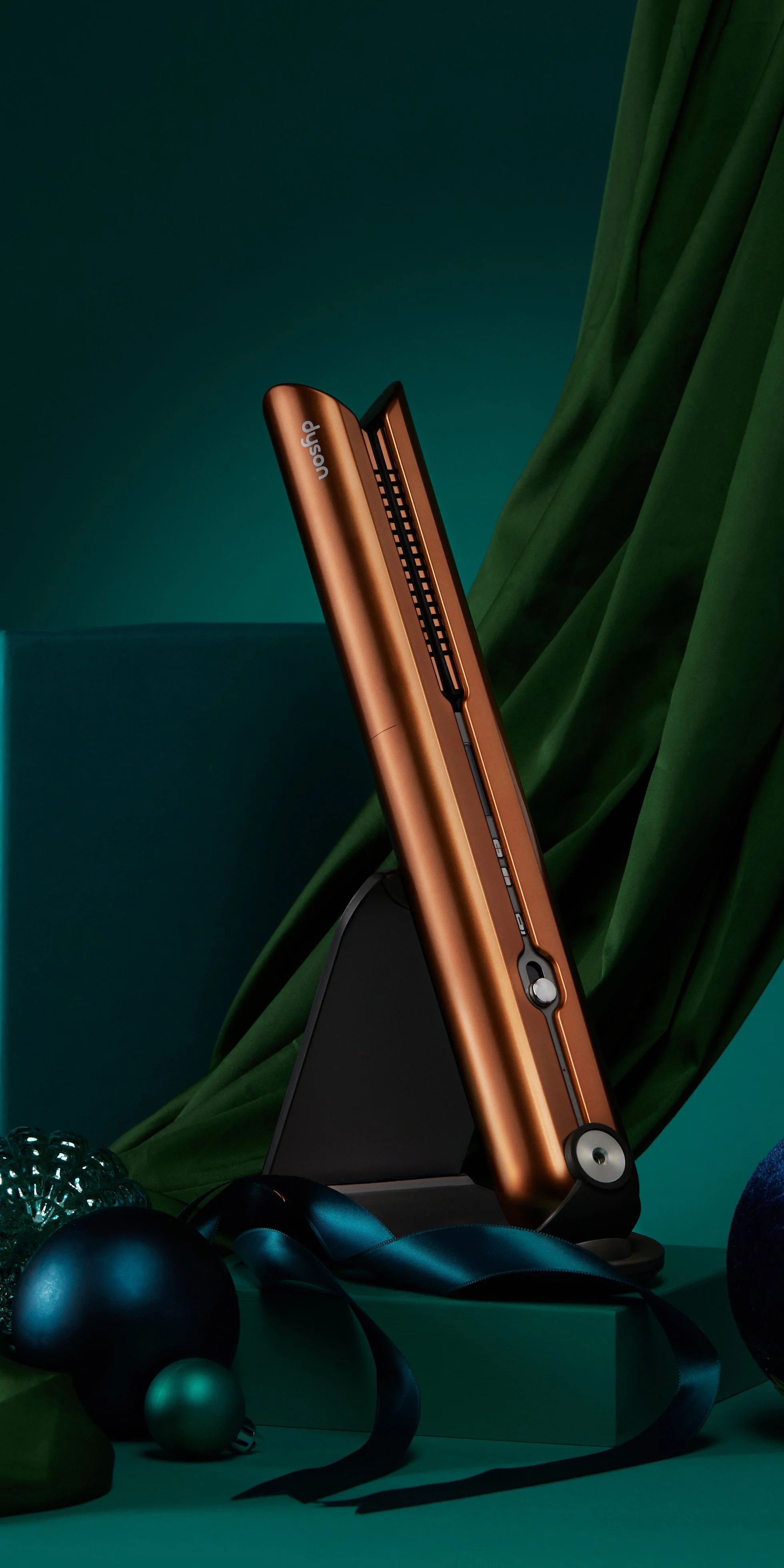 A copper-colored Dyson hair straightener with a black stand, surrounded by dark blue and green decorations and ribbons, set against a teal background with draped green fabric.