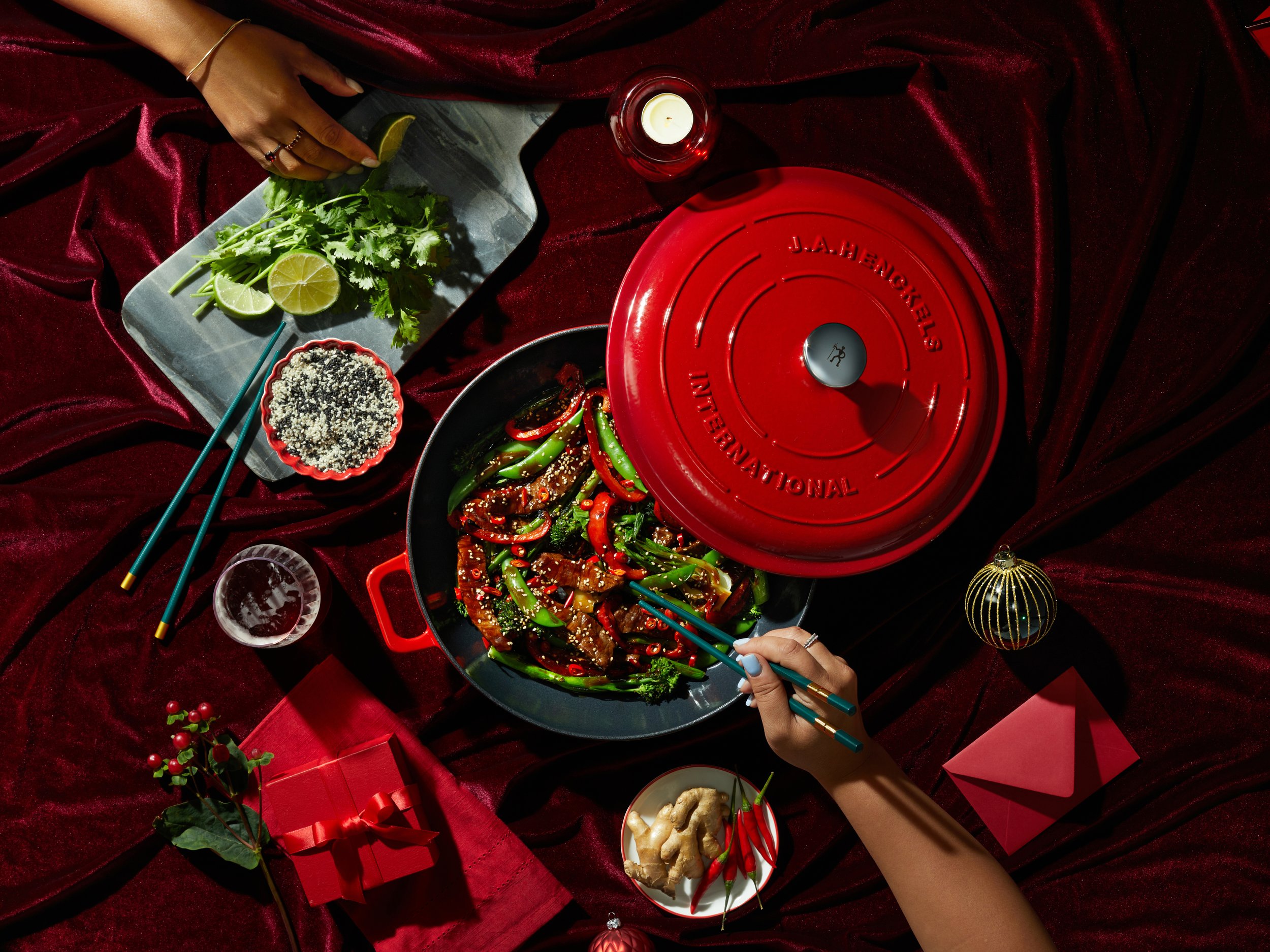 A table with Asian food, including a pan of stir-fry vegetables and meat, a plate of lime and cilantro, ginger, red chili peppers, sesame seeds, and a box of rice, set on a red velvet cloth with chopsticks, a lantern, and decorative items.