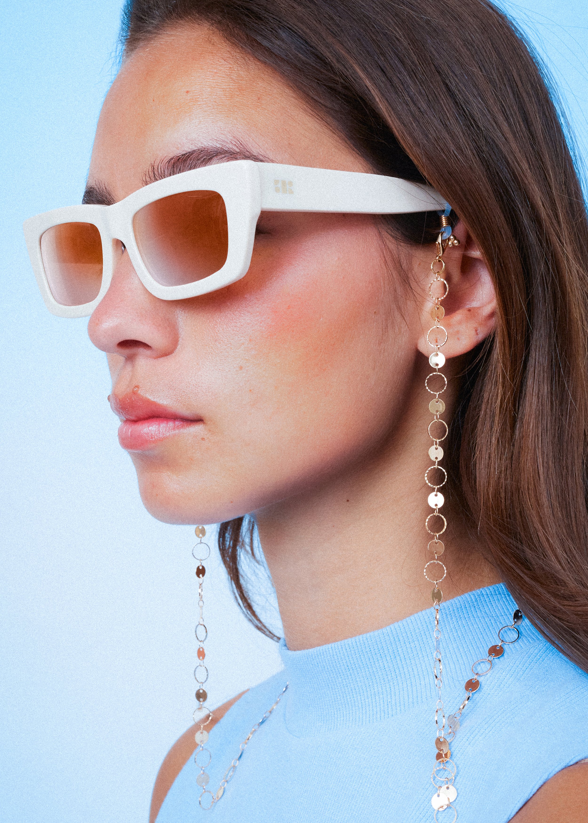 A woman wearing white rectangular sunglasses with brown lenses, a blue sleeveless top, and long gold chain earrings with round and disc-shaped links.