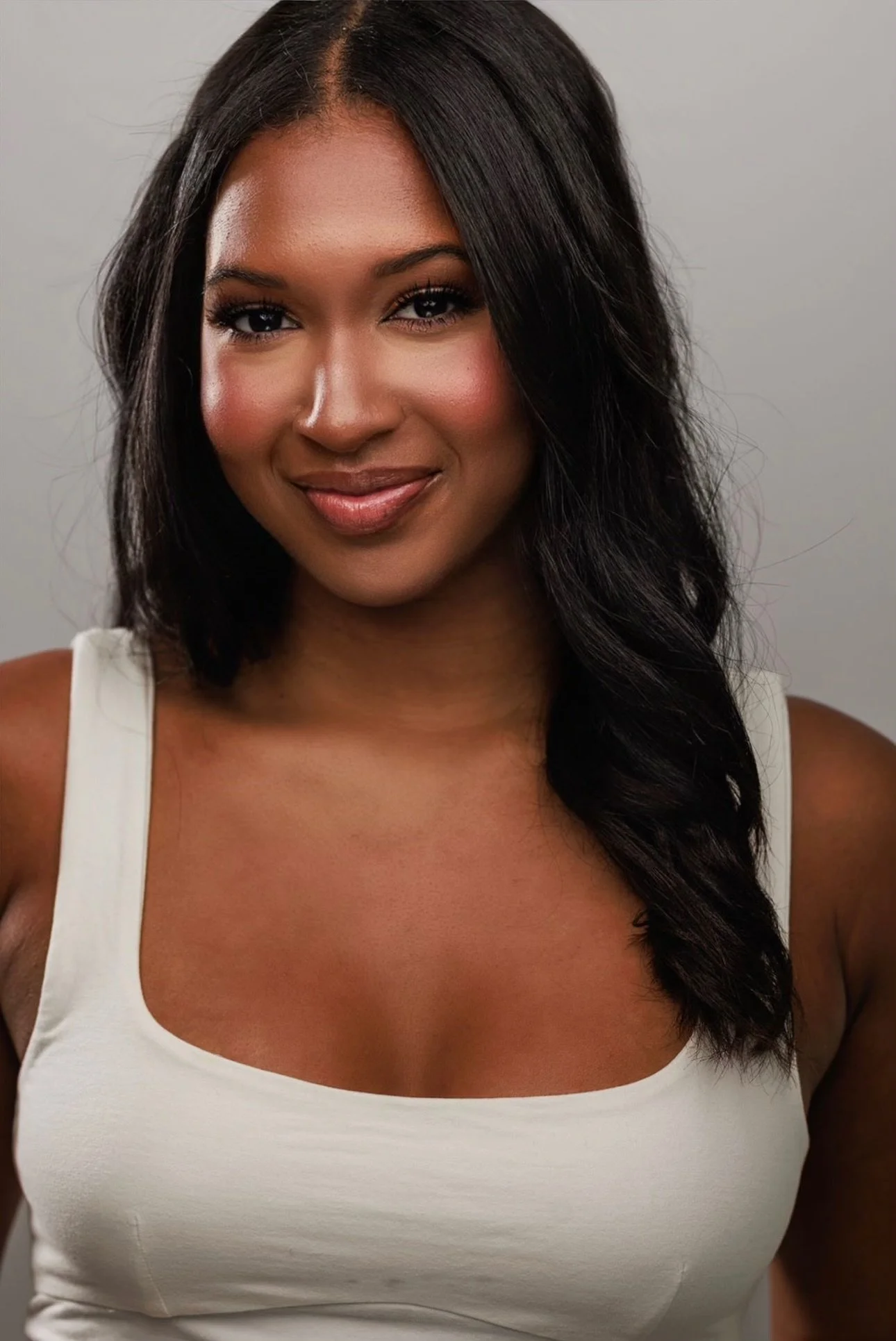 Close-up portrait of a smiling woman with dark, wavy hair wearing a sleeveless white top against a plain gray background.