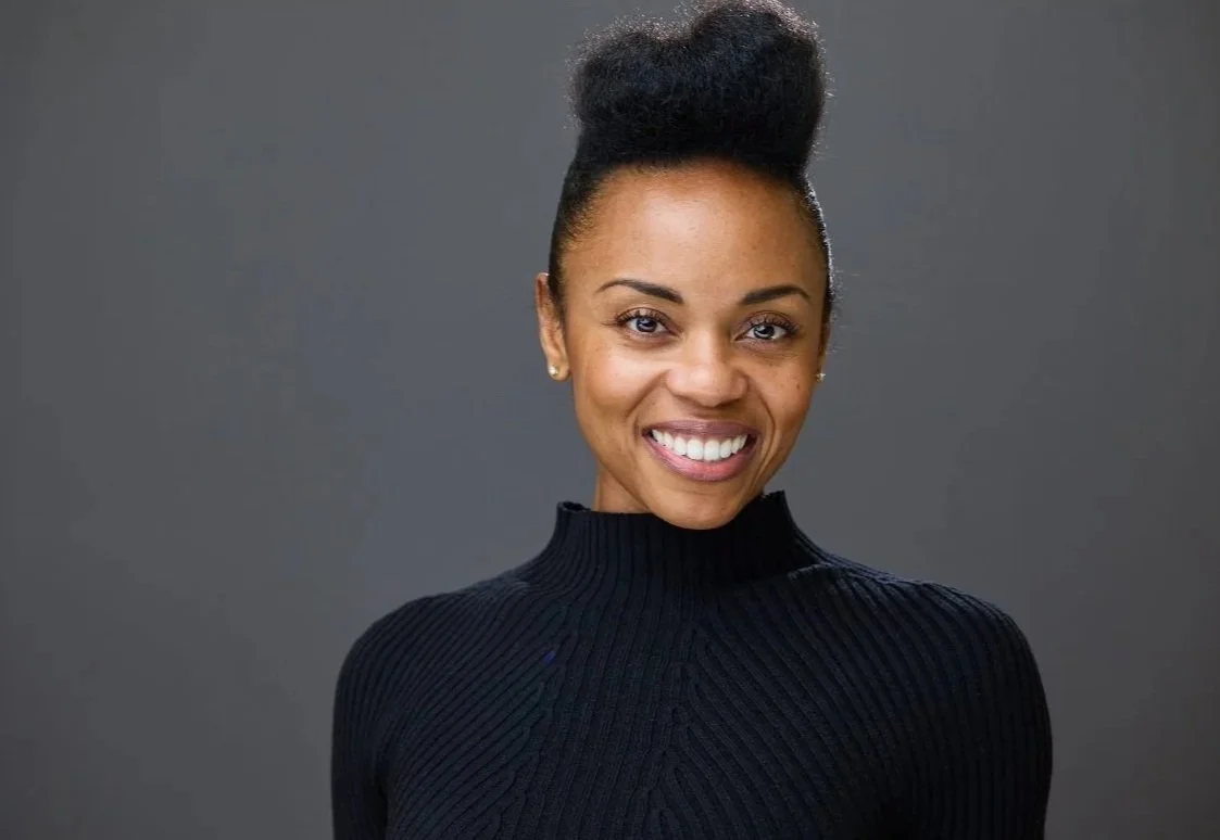 A woman smiling, wearing a black turtleneck sweater, with her hair styled in a high puff, standing against a gray background.