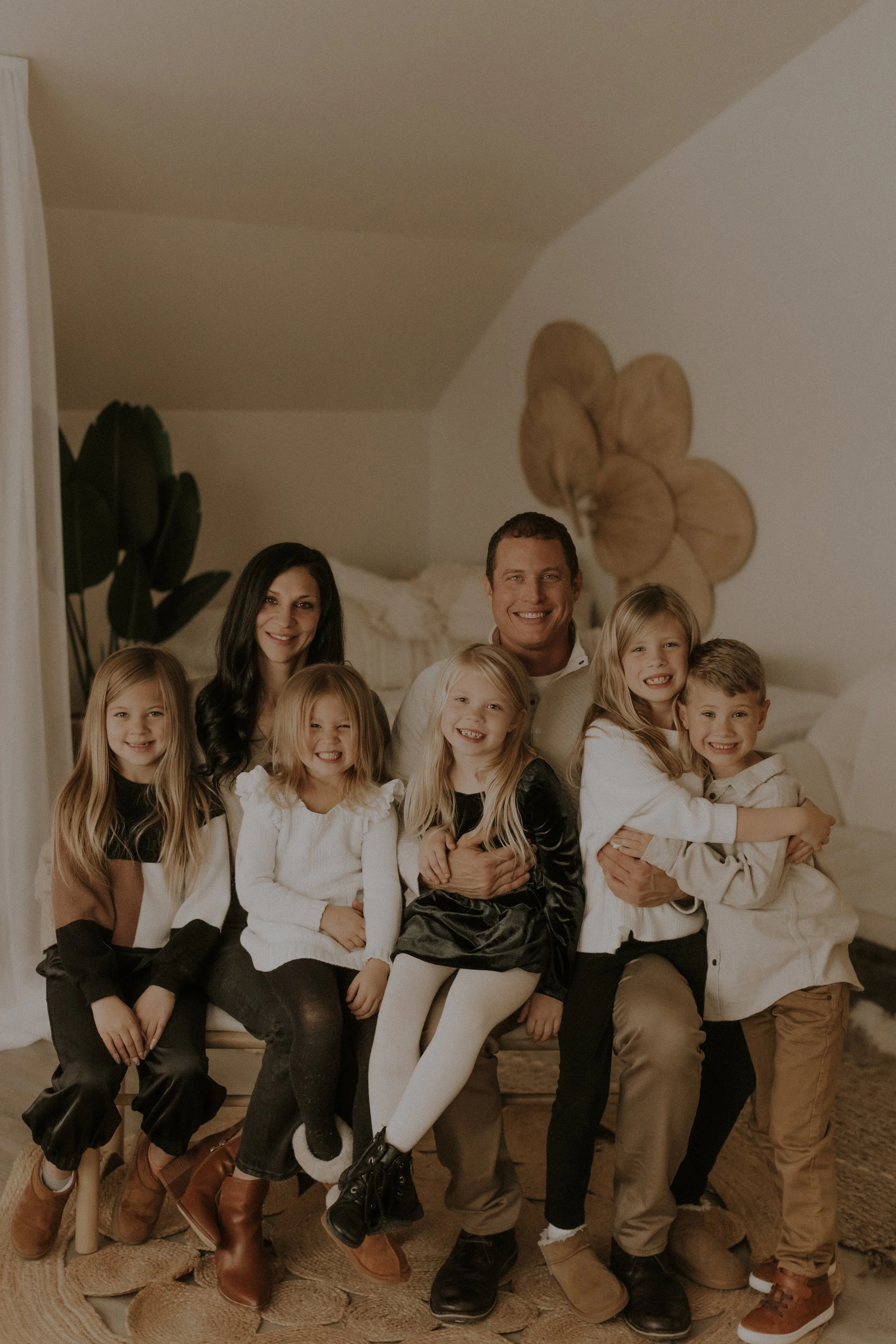 Family portrait of seven people, including two adults and five children, sitting together indoors on a beige rug, smiling and embracing each other.