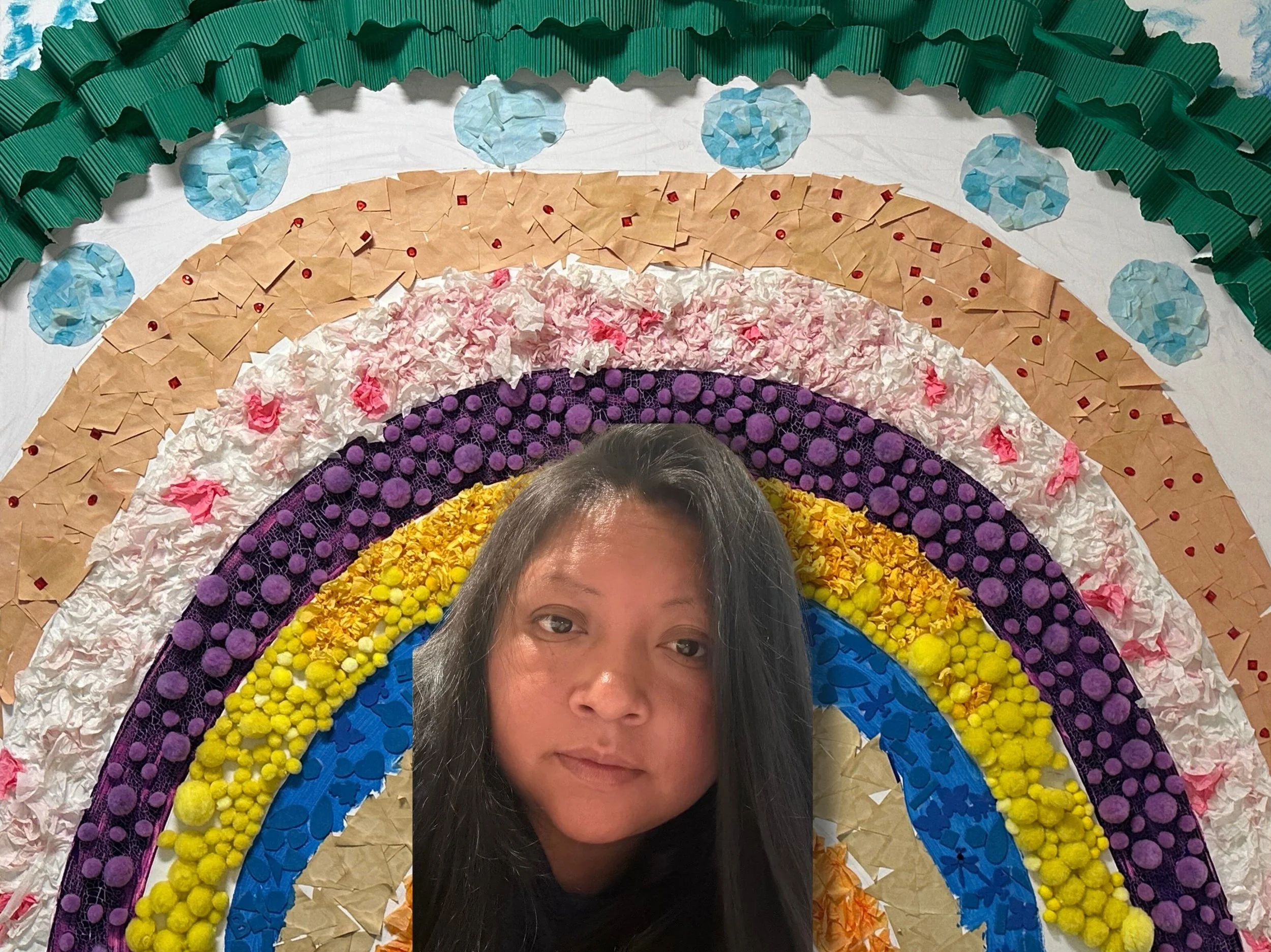 A woman with long dark hair and a neutral expression, standing in front of a colorful rainbow made from various textured and colored paper and materials, including green at the top, followed by light blue, brown, white with pink accents, purple, yellow, and blue. 