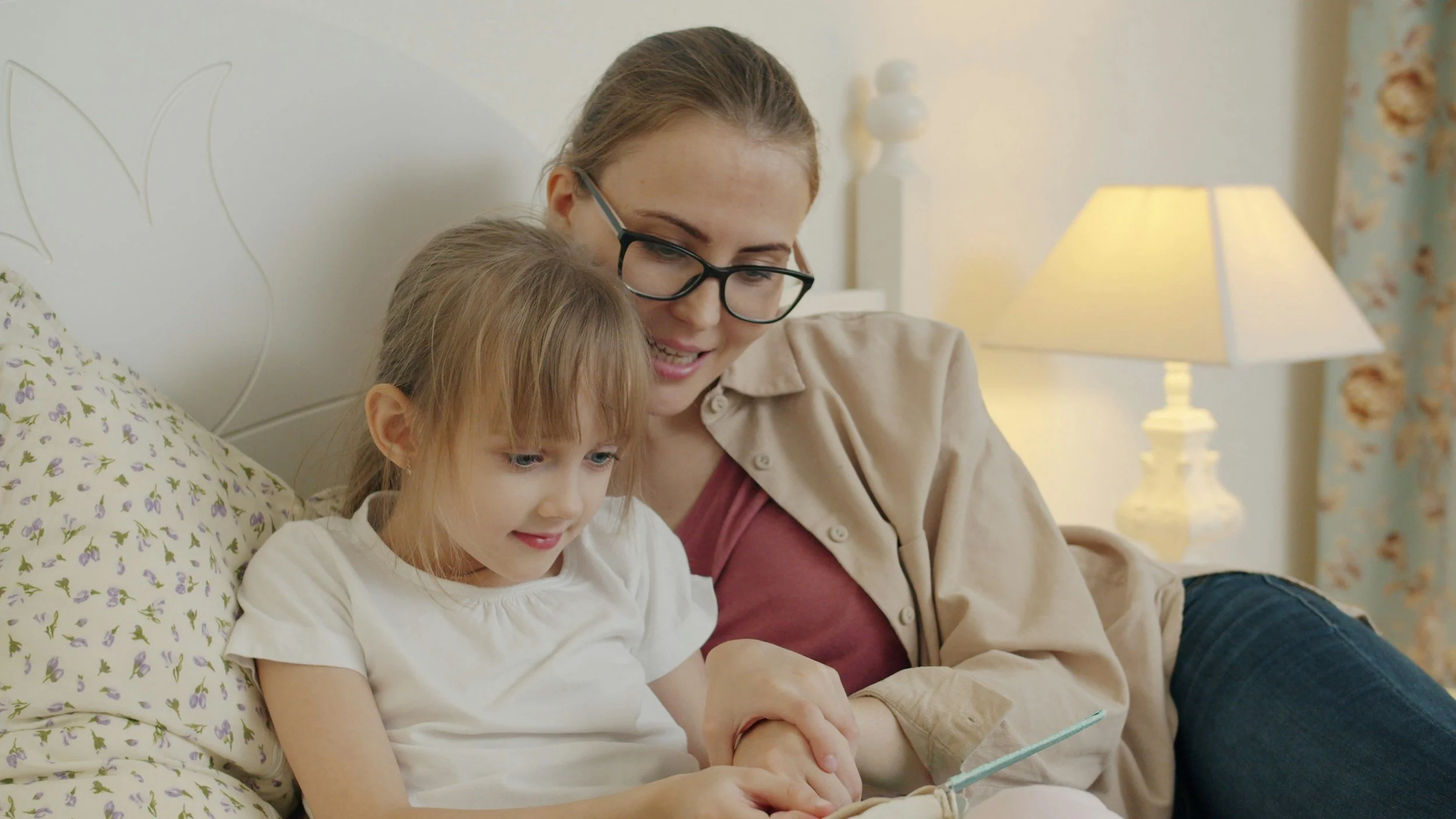 A woman with glasses sitting on a bed with a young girl, both looking at a smartphone together in a warmly lit bedroom.