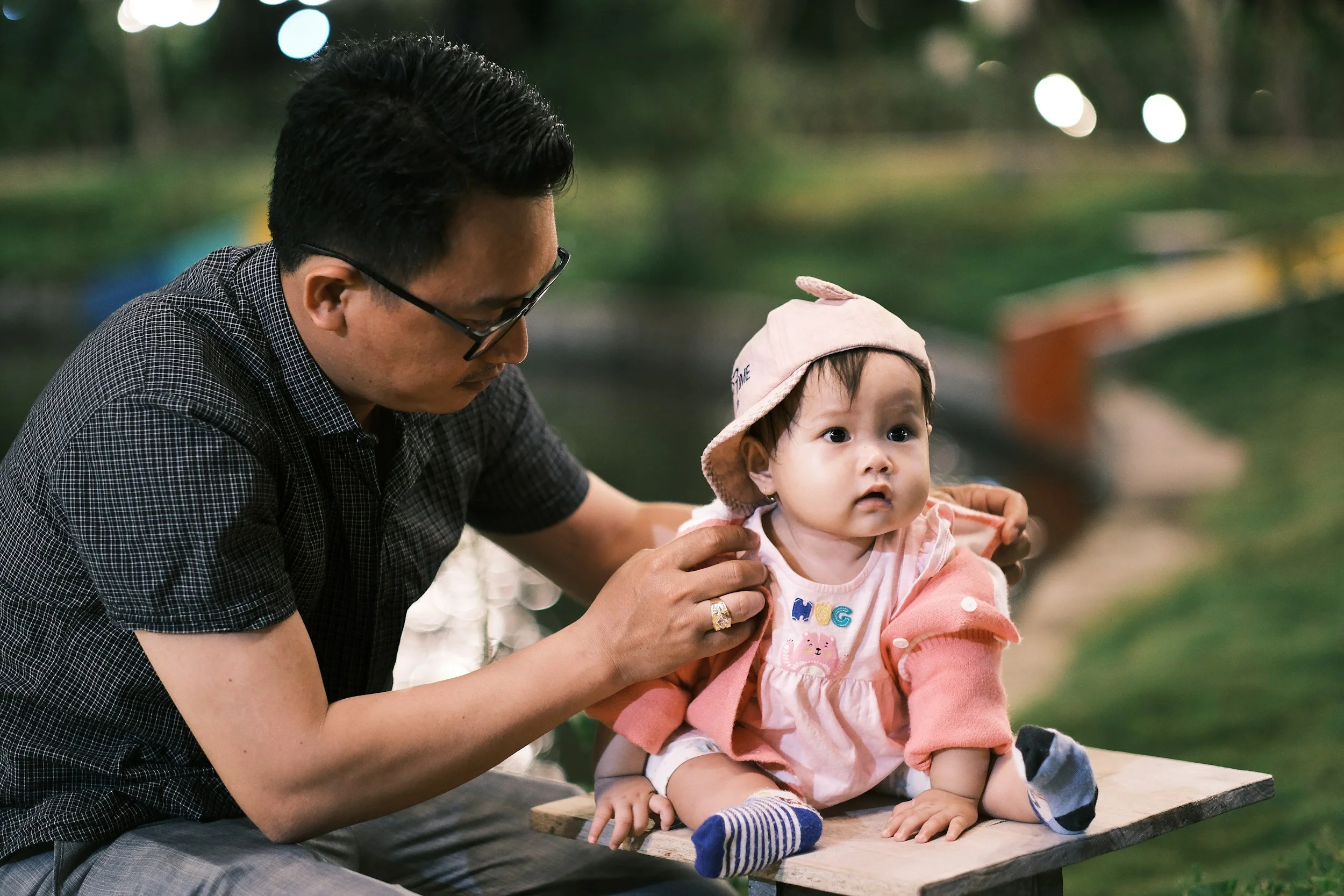 A man is placing an earring on a baby girl sitting on a wooden platform outdoors, with greenery and blurred lights in the background.